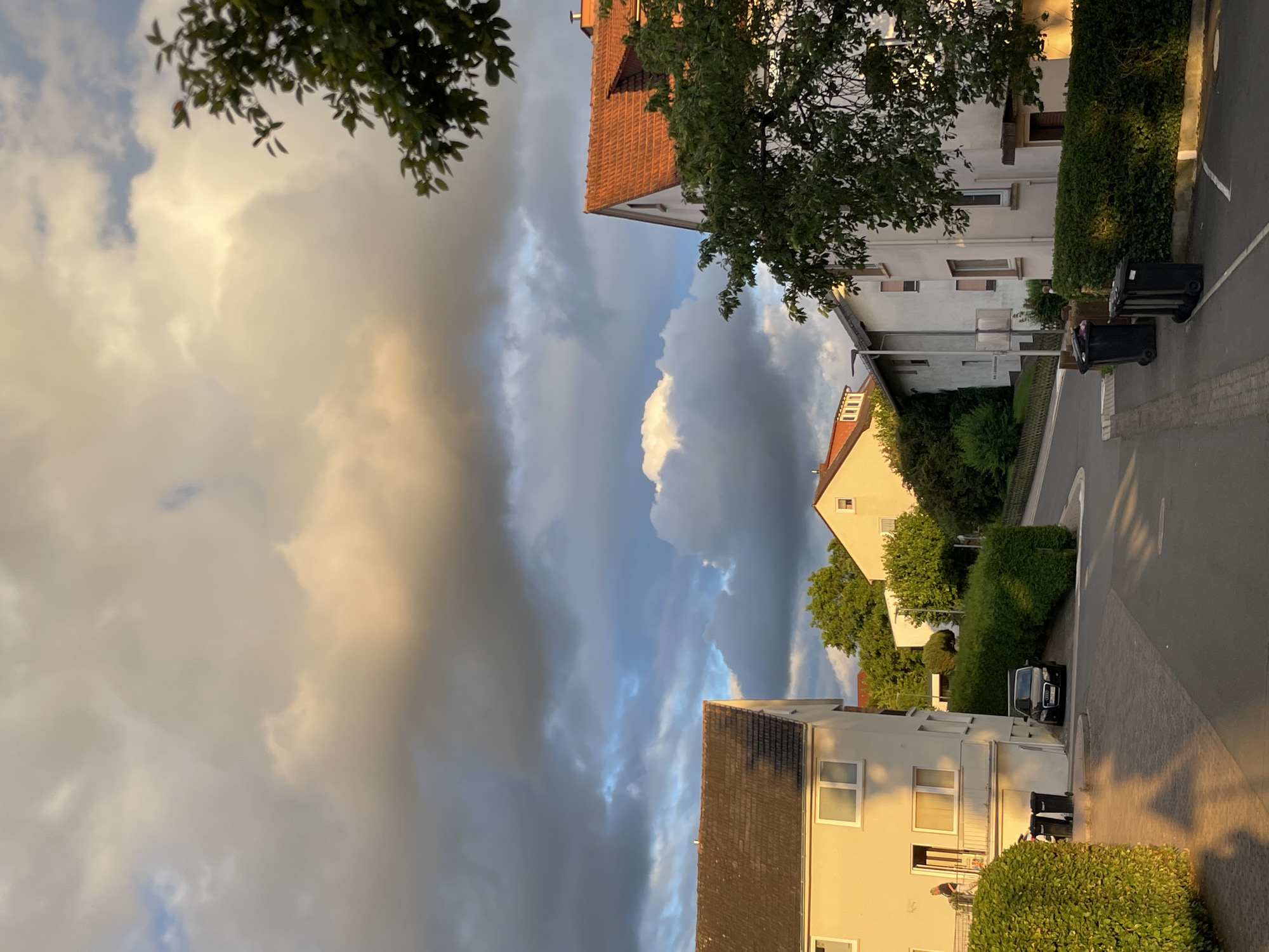 This media file captures a dramatic sky over a residential street in Neukirchen, Germany. The scene is dominated by a striking contrast between luminous, golden-hued clouds on the left and dark, heavy storm clouds with streaks of blue sky on the right. The time of day appears to be late afternoon or early evening, with the warm sunlight illuminating the edges of the clouds and casting long shadows.
Several houses with red-tiled roofs and light-colored facades line the street, interspersed with dense green hedges and trees. The architecture suggests a suburban or semi-urban setting. A black car is parked on the right side of the street, and two black trash bins are visible near the curb. A person is standing on a balcony of one of the houses in the foreground. The overall mood is one of impending change, with the beautiful but threatening sky creating a sense of awe and anticipation. There is no visible text in the image.
