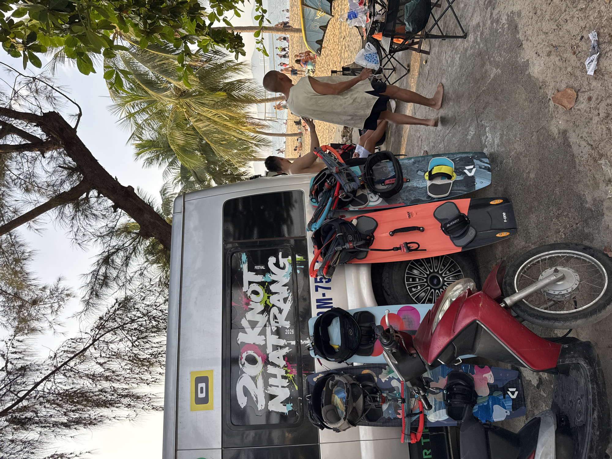 The image depicts a scene on a beach in Nha Trang, Vietnam, likely during an event related to kitesurfing or watersports, given the presence of kiteboards, harnesses, and a van with the text "20 KNOT NHATRANG FESTIVAL 2026". The van is parked on a paved area adjacent to a sandy beach where several people are visible.
In the foreground, a red scooter is parked on the left. To the right of the scooter, leaning against the van, are three kiteboards with their associated harnesses. The van itself is silver and has a large graphic on its rear window.
Two men are standing near the van. One man, wearing a light-colored sleeveless shirt and dark shorts, is looking towards the beach and appears to be interacting with the other man, who is partially visible and shirtless. Further back on the beach, several groups of people are seen relaxing, playing volleyball, and engaging in other beach activities. Palm trees and foliage frame the scene, suggesting a tropical environment. The sky is overcast with a hint of sunlight. A table with chairs and some supplies is set up on the right side of the paved area.