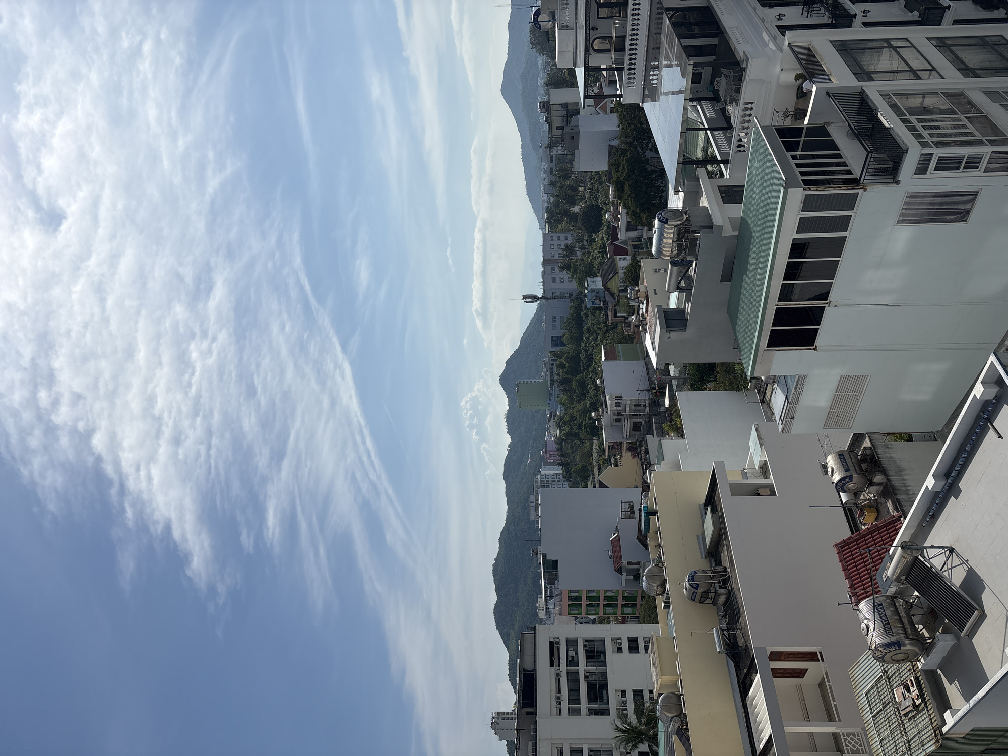 This is a high-angle, daytime view of Nha Trang, Vietnam, looking from a building across the city. Mountains are visible in the distance, separating the sky and the urban landscape. The sky is partially cloudy, showing a mix of blue and wispy white cloud formations. In the foreground, are the rooftops of multi-story buildings, some with solar water heaters. Vegetation is interspersed among the buildings, with denser patches closer to the base of the mountains. The orientation of the image is rotated 90 degrees counterclockwise.