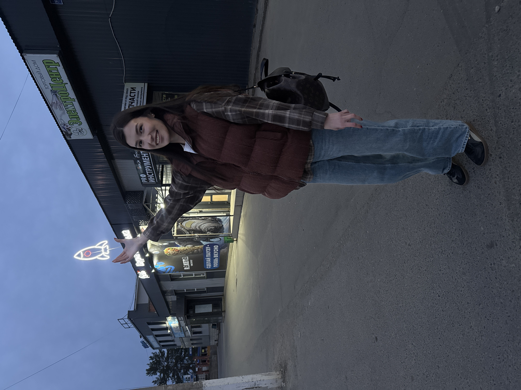 The image captures a woman outdoors in Chaikovsky, Russia, against an urban backdrop. She is wearing a plaid shirt, a brown vest, jeans, and black shoes. She is smiling and holding out her hand towards a neon sign shaped like a rocket, which is mounted on a building. A backpack is visible on her shoulder. In the background are commercial buildings with signs, including one that reads "Elektrotsentr" (Электроцентр) and another for "Raketo" (РАКЕТО) шаурма. Streetlights illuminate the scene, and the sky suggests it is dusk or early evening.