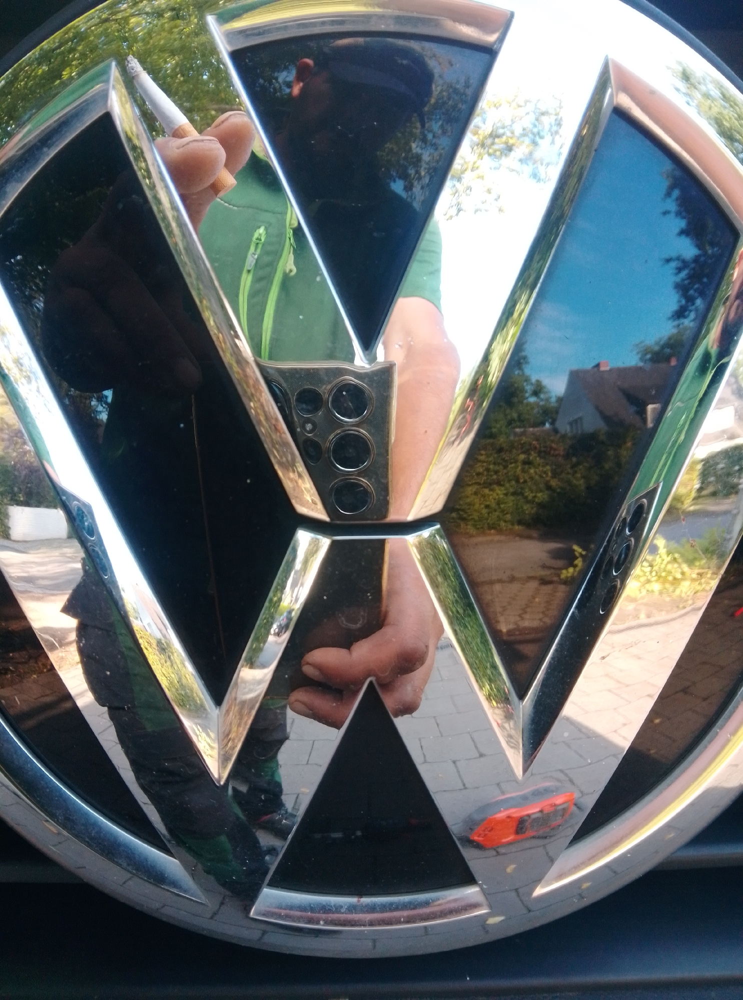 The image shows a close-up reflection of a person in the chrome and black emblem of a Volkswagen car. The person is holding a lit cigarette in their right hand, with their fingers visible against the shiny surface. They are wearing a green jacket and dark pants, with a reflection of their lower body visible. The background reflection shows trees, a house with a red roof, and a paved driveway, suggesting an outdoor setting. The car emblem is shiny and reflects the surroundings with a slight distortion. There is no explicit indication of the specific location within Hamburg, Germany.