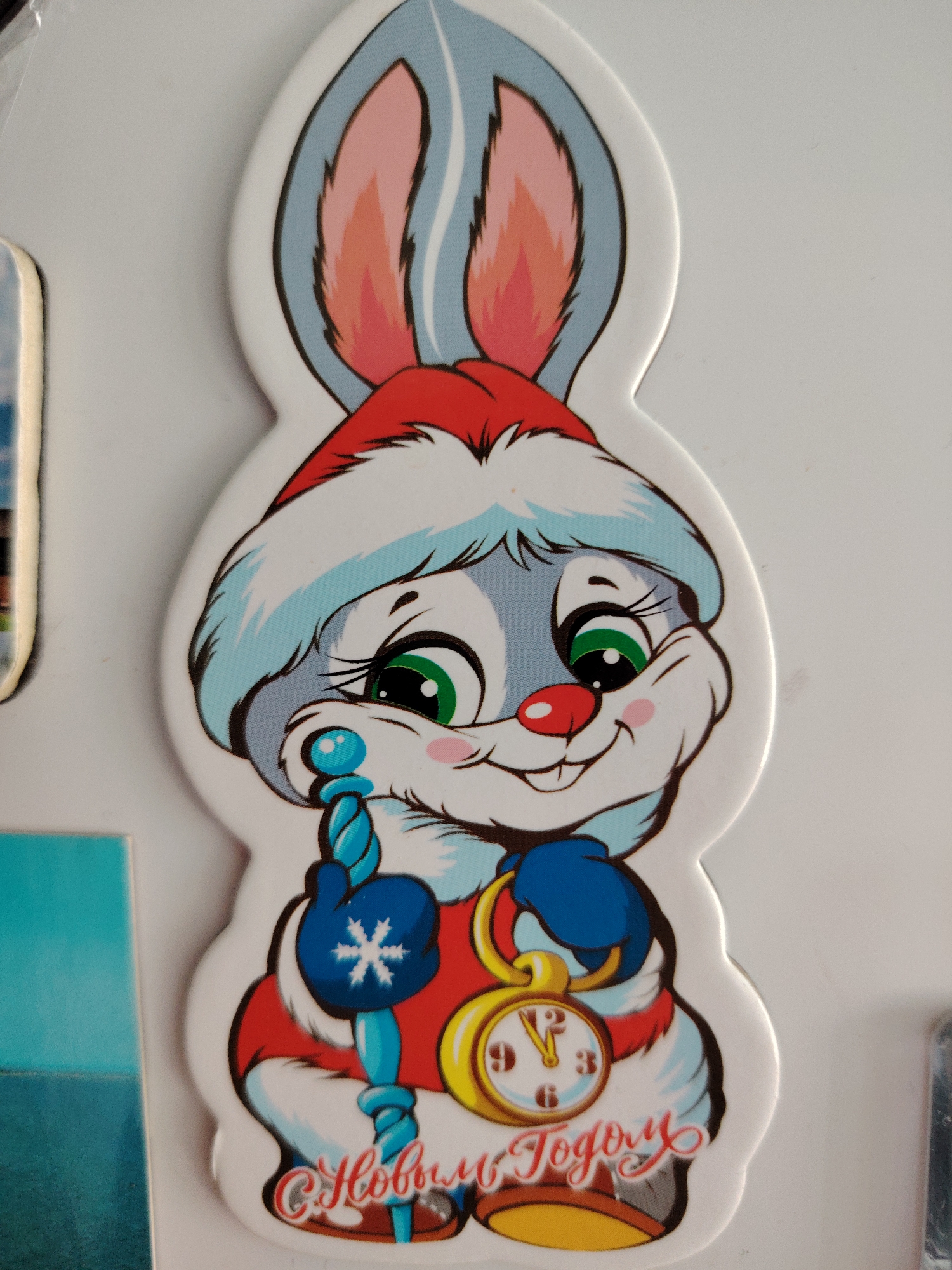 The image features a close-up of a festive, cartoon-style rabbit character, likely a magnet or sticker, set against a plain white background. The rabbit is designed with predominantly white and light blue fur, sporting large, expressive green eyes with long eyelashes, a small red nose, and rosy pink cheeks. Its inner ears are also pink.
The character is adorned in winter holiday attire, including a red and white Santa hat. It wears blue mittens, one of which has a white snowflake emblem. In its left mittened paw, the rabbit holds a light blue staff or scepter with a spiraled design and a spherical top. In its right mittened paw, it clutches a golden pocket watch with a white face and black numerals, indicating the time is set just past midnight. The rabbit has a wide, happy smile, suggesting a joyful demeanor.
Below the rabbit, in decorative red script, the text "С Новым Годом" is visible, which translates from Russian as "Happy New Year." The overall scene portrays a celebratory atmosphere, likely associated with the New Year holiday, given the symbols of the Santa hat, snowflake, and the "Happy New Year" greeting. The location context for this media file is Сусады-Эбалак, Russia.