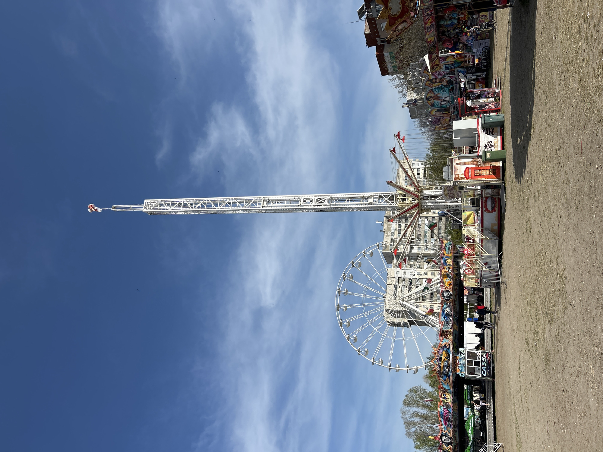 The image captures an outdoor amusement park or fairground scene under a clear, bright blue sky with sparse white clouds. Dominating the left side of the frame is a tall, white metal drop tower ride, its structure extending vertically towards the sky, with a ride carriage visible near its apex. To its right, a large white Ferris wheel stands prominent, featuring numerous passenger gondolas.
In the mid-ground and extending to the right, a variety of colorful fair stalls and smaller rides are visible. These booths are adorned with vibrant decorations, some appearing to offer games or prizes. Notable among them is a distinct red booth resembling a British telephone box, and several green trash cans are placed nearby. The ground beneath the attractions is unpaved, consisting of dirt or gravel.
In the background, behind the rides and stalls, several multi-story buildings can be seen, indicating an urban or semi-urban setting, consistent with the location context of Plovdiv, Bulgaria. Trees are also visible towards the lower right. Several individuals are scattered near the bases of the rides and stalls, suggesting the fair is operational. The word "CASSA" is visible on one of the booths near the Ferris wheel, signifying a ticket office or cashier station. The scene is brightly illuminated by daylight, indicating a sunny day.