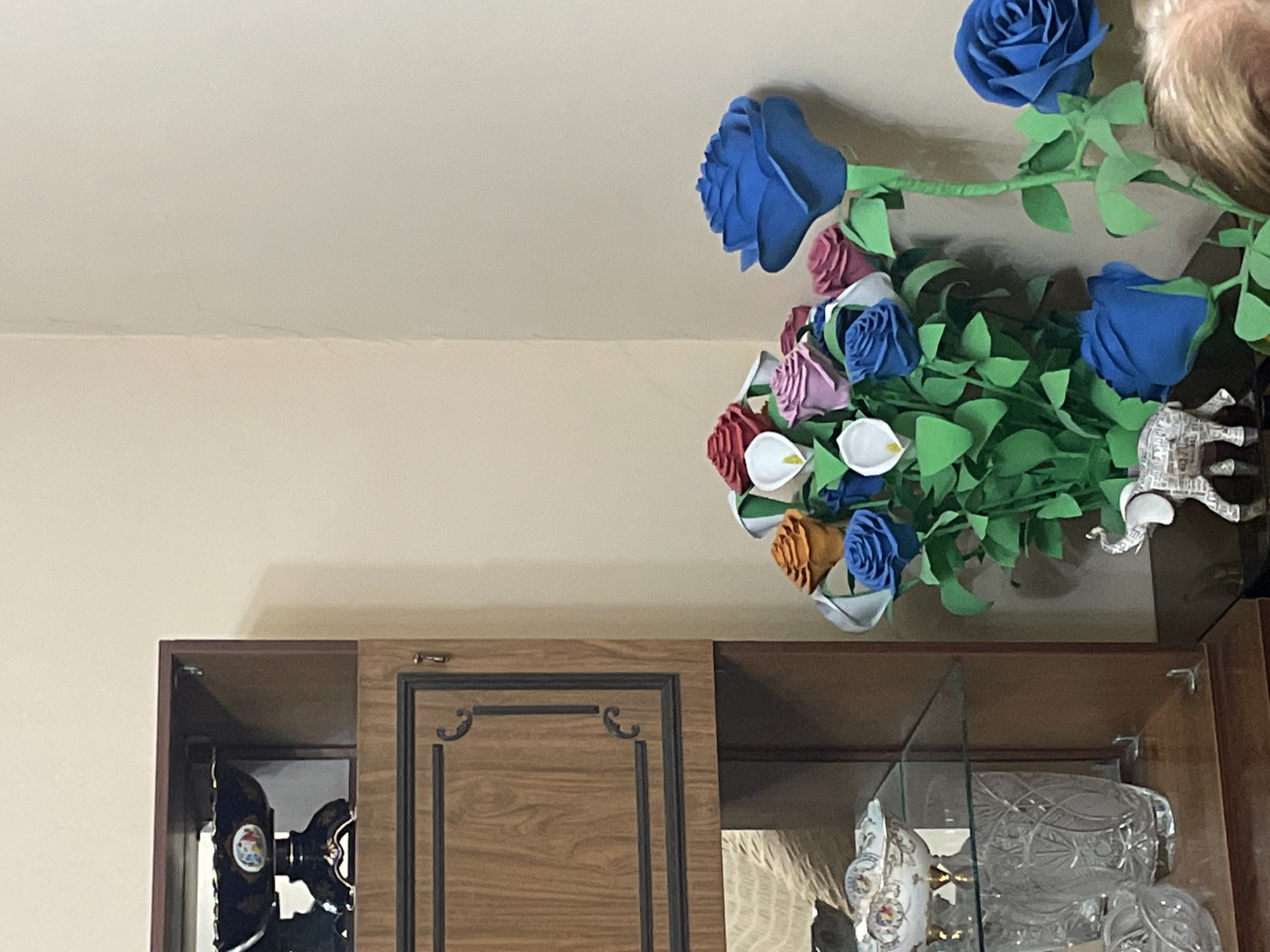 The image shows a corner of a room, likely indoors, with a cabinet and decorative items. The cabinet is made of wood, with one section closed and the other open, revealing items inside. Inside the open section, there's what appears to be a sewing machine on one side and decorative glass items on the other. Above the cabinet sits a colorful bouquet of artificial flowers in shades of blue, pink, white, and orange. There's also a small decorative elephant figure placed nearby. The background consists of a plain off-white wall and ceiling. No people are clearly visible. Without additional information, the location being Yerevan, Armenia cannot be confirmed based solely on this image.