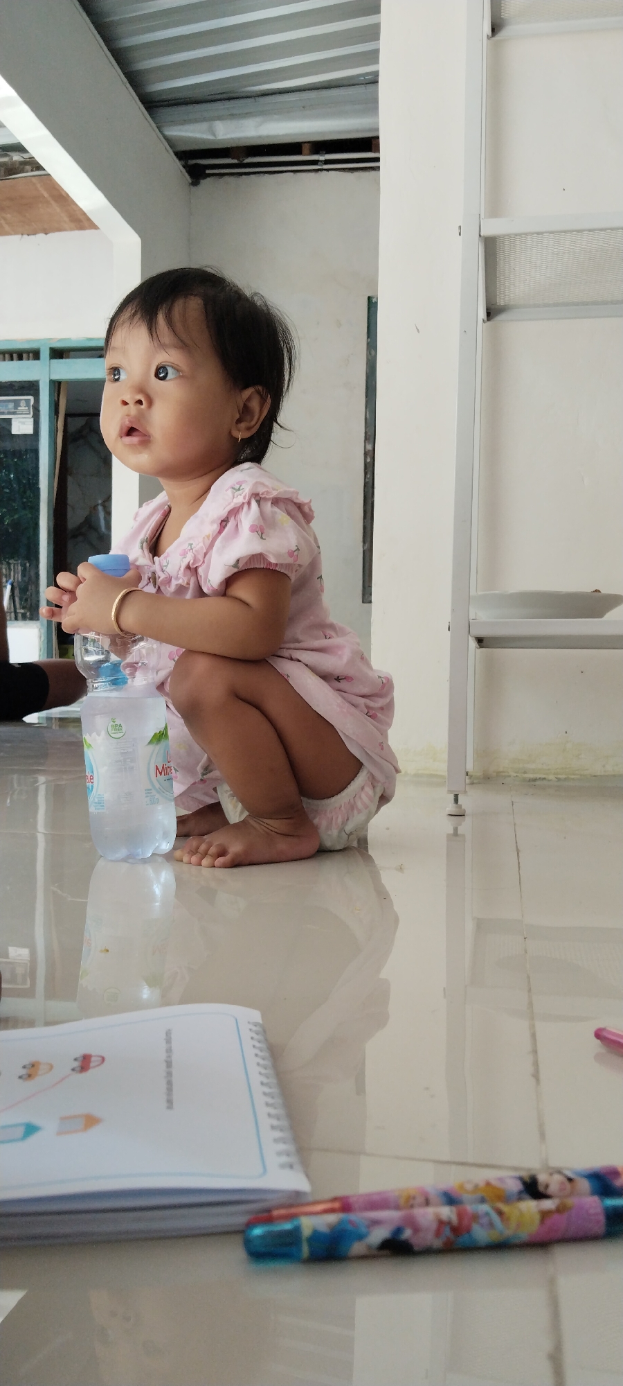 A young Asian child is squatting on a shiny, tiled floor, holding a plastic bottle of water. The child, wearing a pink dress with cherry motifs and a diaper, looks to their left with wide eyes. A golden bracelet is on their right wrist. The environment appears to be an indoor or semi-outdoor space with a corrugated metal roof visible above, and white walls and shelving units in the background. A notebook with simple drawings is on the floor in the foreground, along with two pens decorated with cartoon characters. The lighting is natural and soft. The scene suggests a casual, domestic setting, possibly in Kecamatan Pati, Indonesia, given the provided location context.