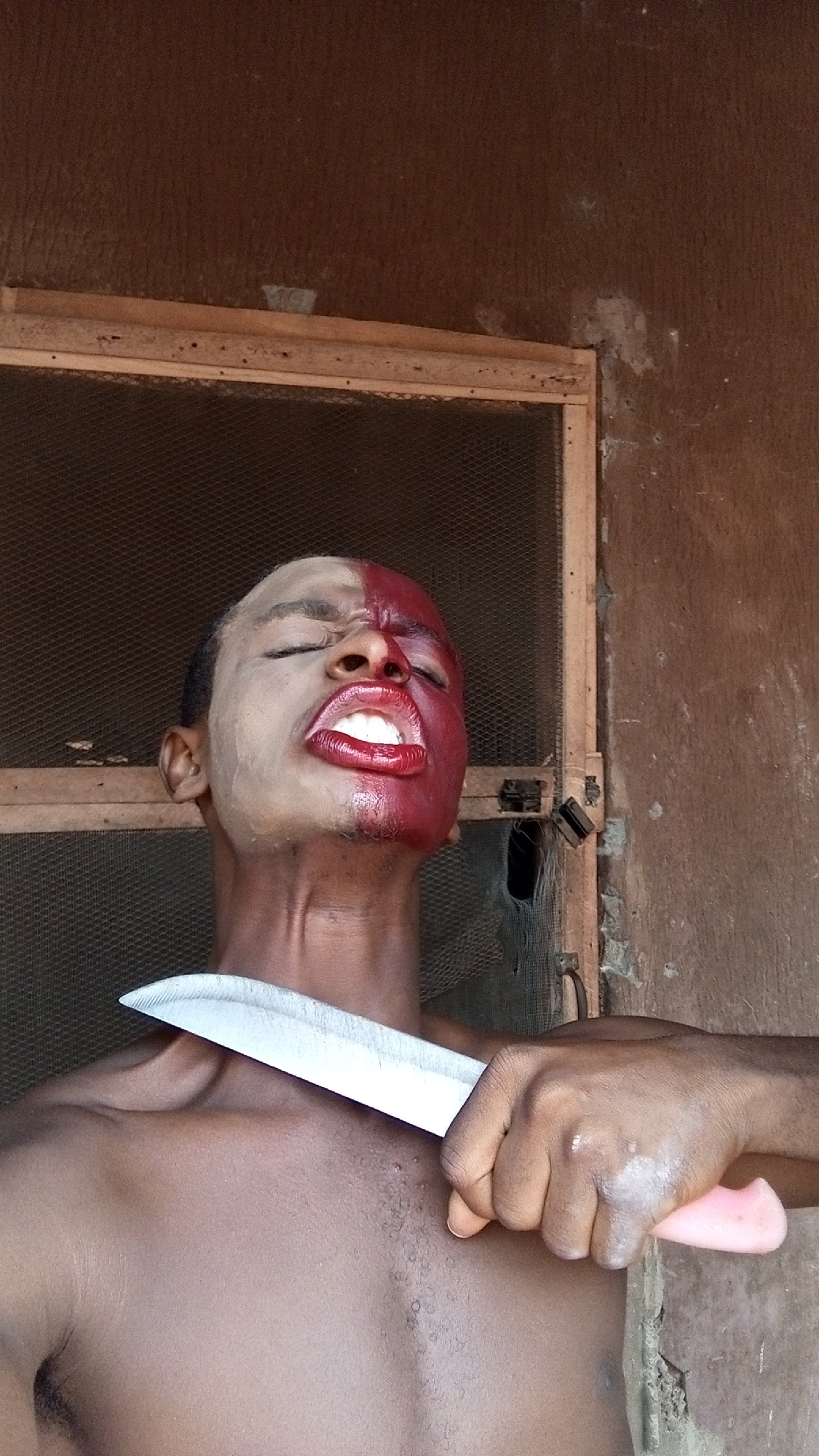 A shirtless person with one half of their face painted red and the other a lighter shade holds a knife to their neck, with their eyes closed and mouth open. The backdrop is a window screen and a weathered wall. The location is Unknown, Unknown.