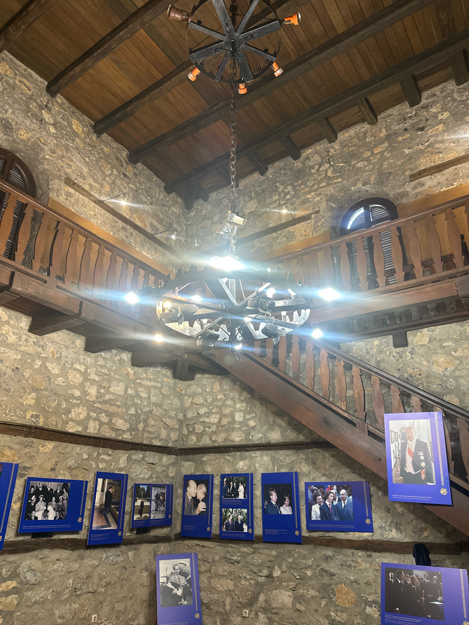 The image depicts the interior of the Royal Complex in Topola Municipality, Serbia. The scene is a stone-walled hall with wooden beams and a wooden balcony and staircase. A large, ornate metal chandelier hangs from the ceiling, illuminated by spotlights. Several framed photographs are displayed on the stone walls, featuring formal portraits of individuals in what appear to be historical or ceremonial attire. The photographs are mounted on blue backing with gold trim. The overall impression is of a historical exhibit or museum space within the complex.