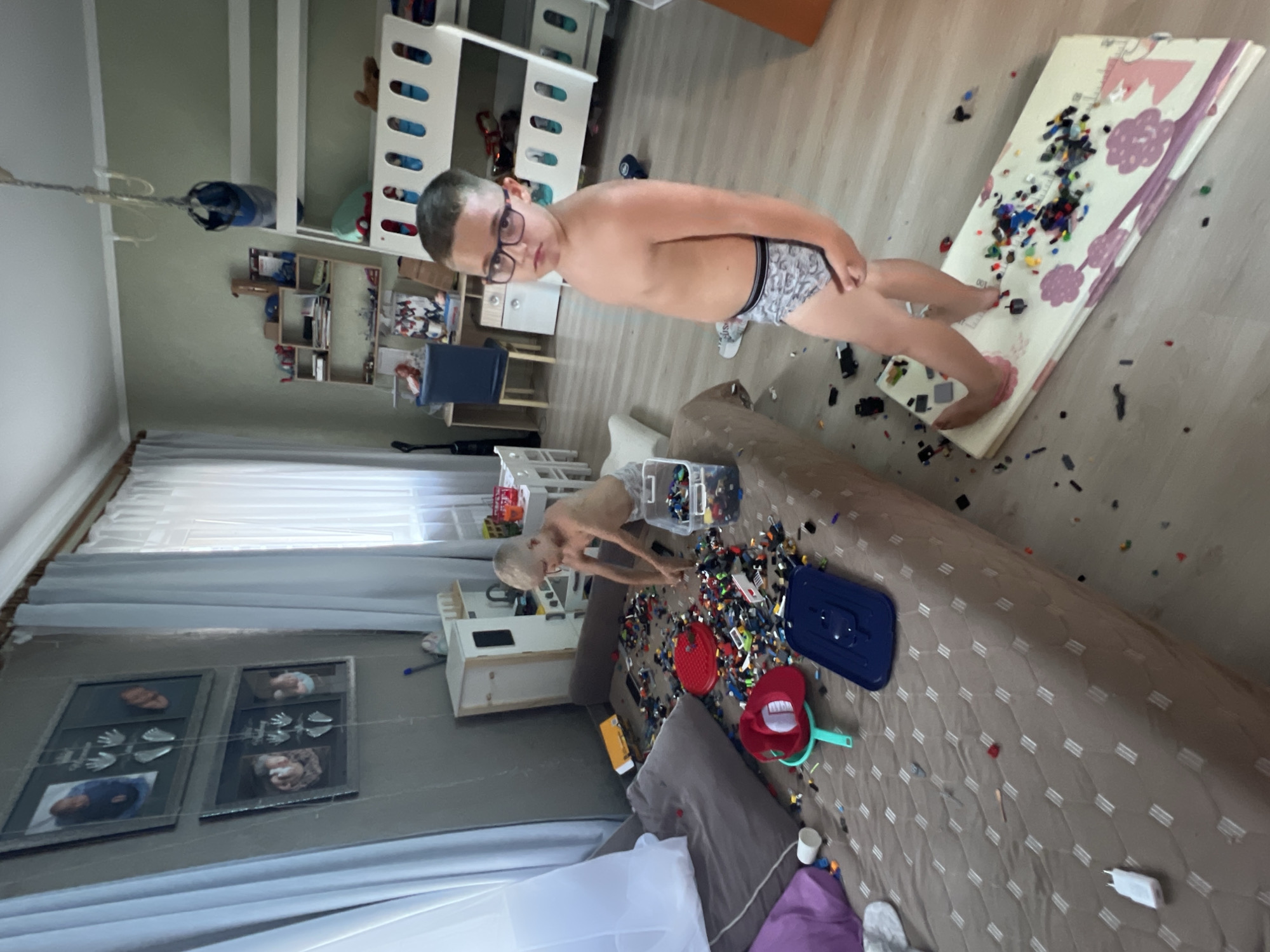 An indoor scene in what appears to be a child's bedroom or playroom in Sochi, Russia, features two young boys amidst scattered Lego bricks. The room has light green-gray walls and light wood-colored flooring.
In the foreground to the right, a boy with short, dark hair and eyeglasses stands facing the camera. He is shirtless, wearing patterned underwear, and is barefoot, with one foot partially on a folded white and pink play mat that is covered in numerous multi-colored Lego bricks.
In the mid-ground to the left, a younger boy with short, blonde hair is positioned on a brown quilted daybed or large mattress. He is shirtless and wearing light-colored underwear, actively engaged in playing with or sorting a large quantity of multi-colored Lego bricks that are spread across the bed. A clear plastic container of Lego is visible next to him. Other items on the bed include a dark gray pillow, a purple pillow, a white charging adapter, a red plastic bowl or hat, a teal scoop or strainer, and a dark blue plastic tray or lid.
The left wall features two framed displays, each containing an infant's photograph, handprints, and footprints. A large window with sheer white and light blue curtains is visible on the far left.
In the background, a white bunk bed occupies the right side of the room. Above it, a multi-pronged ceiling fixture hangs, with a blue baseball cap suspended from it. A wooden shelving unit filled with various items is situated against the back wall, and a small desk with a blue chair is partially visible below it. Further to the left in the background, a white wooden play kitchen or cabinet is present, along with a small white table and two chairs.