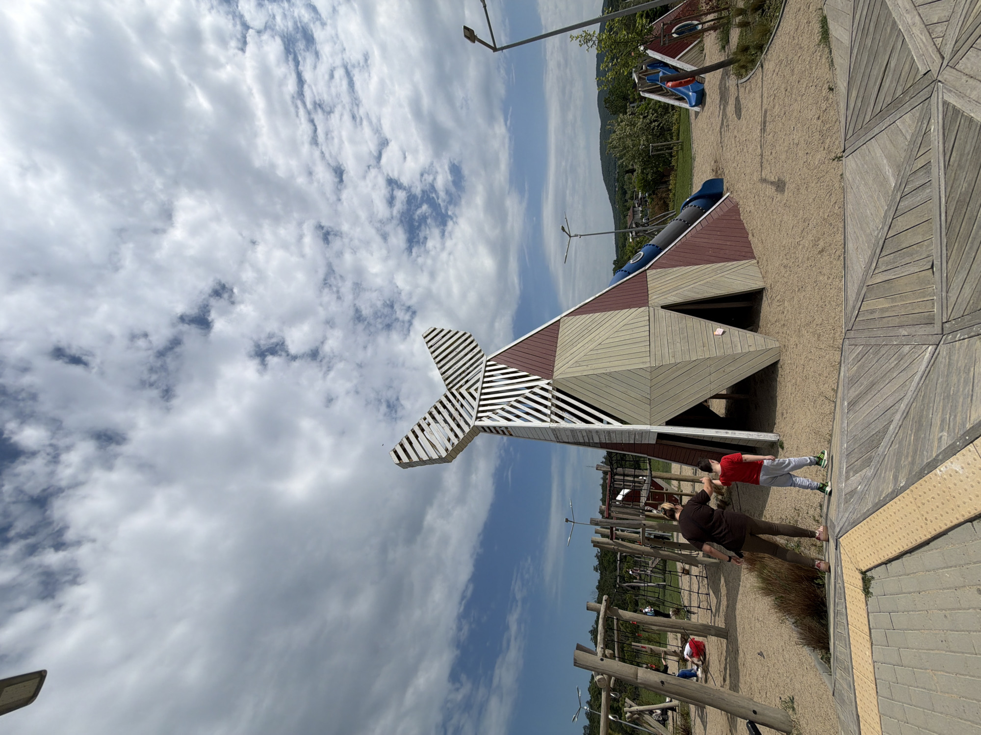 The image shows a playground in Burgas, Bulgaria, featuring a wooden structure resembling a stylized whale or spaceship. The structure has a slide extending from one side. Two individuals are present on the right side of the frame. One is an adult wearing a dark shirt and brown pants, and the other is a child wearing a red shirt and gray pants. They are standing near a wooden walkway. In the background, there are more playground structures and trees. The sky is partially cloudy.