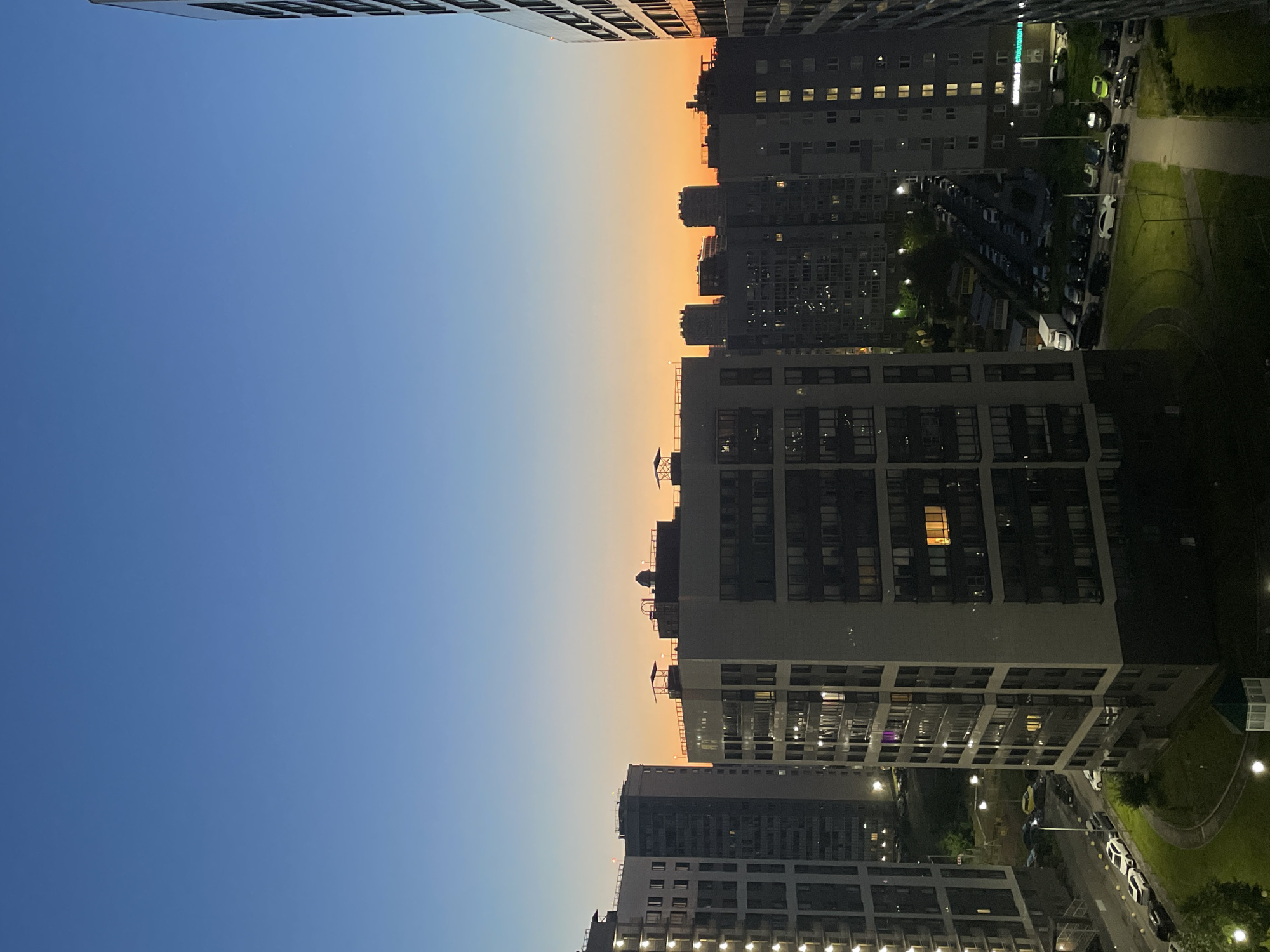 The image depicts a twilight view of a modern residential district in Sinyavino, Russia. A gradient sky transitions from a deep blue at the top to a vibrant orange glow near the horizon, suggesting either sunrise or sunset. Silhouetted against this sky are numerous multi-story apartment buildings, characterized by their uniform window patterns and varied architectural designs. Some buildings have lights illuminated within, indicating occupancy.
In the foreground and midground, several of these apartment buildings are clearly visible. The building closest to the viewer's right edge is tall and modern with a dark facade and numerous windows. To its left, a lighter-colored, complex apartment building dominates the center of the frame, its many windows reflecting the ambient light. Further to the left, another tall, light-colored apartment building with a repeating facade is seen. In the background, a dense cluster of apartment buildings forms the city skyline, fading into the distance.
The ground level of the residential area shows paved roads with parked cars. Multiple vehicles, including sedans and SUVs of various colors, are arranged in parking lots and along the streets. Green spaces, likely lawns or small park areas, are interspersed between the buildings and roads. Streetlights cast pools of light, illuminating parts of the pavement and grassy areas. The overall scene is one of urban development and the evening or early morning transition in a contemporary Russian city. There are no discernible people or interactions in the image.