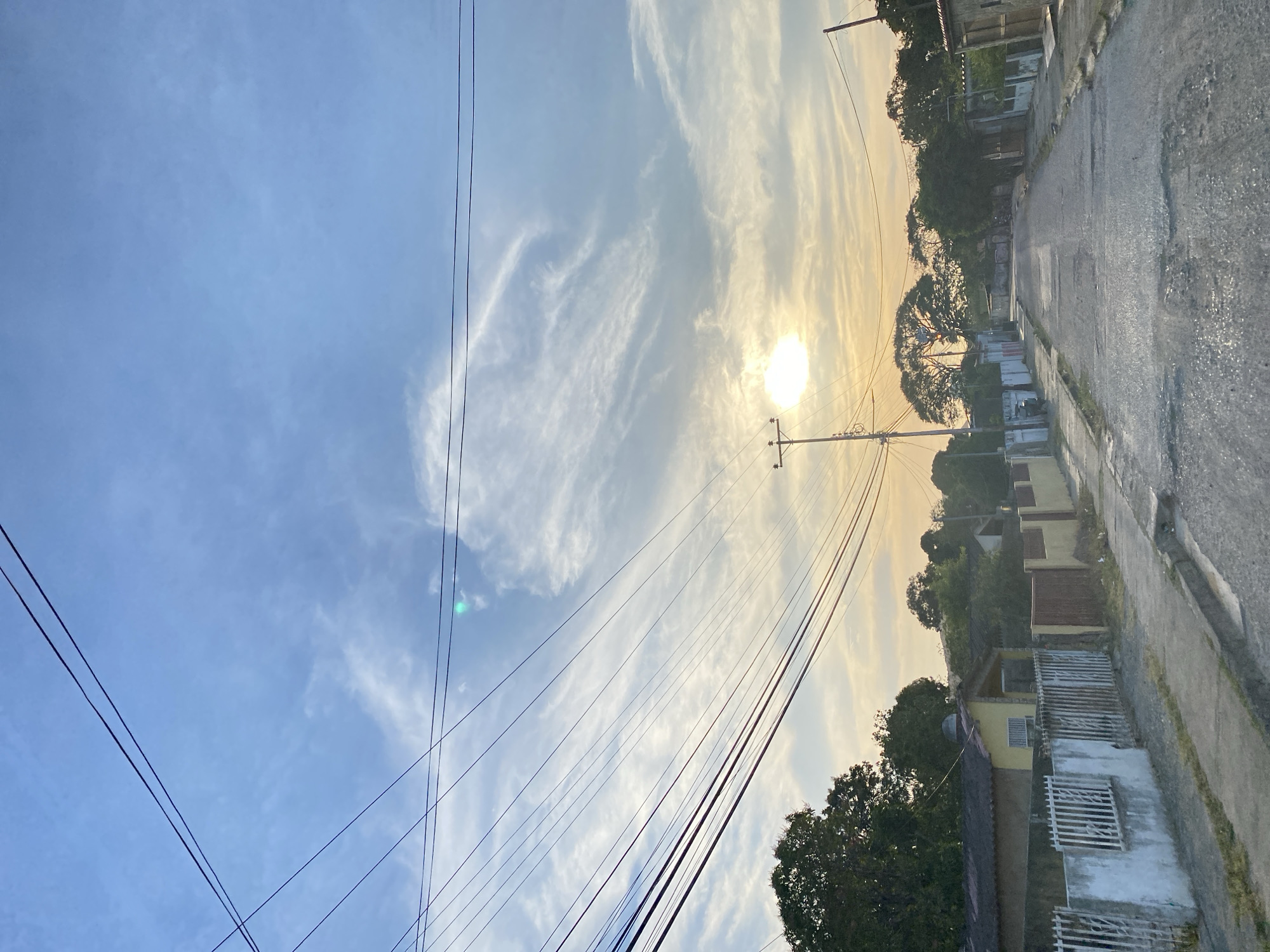 The image depicts a street in Juan Griego, Venezuela, at either sunrise or sunset. The sun is visible through thin clouds, positioned low in the sky. A row of low-rise buildings lines the right side of the street. Utility poles with numerous wires traverse the sky. The pavement appears wet or damp. The overall view is captured at an oblique angle. No people are visible in the shot.