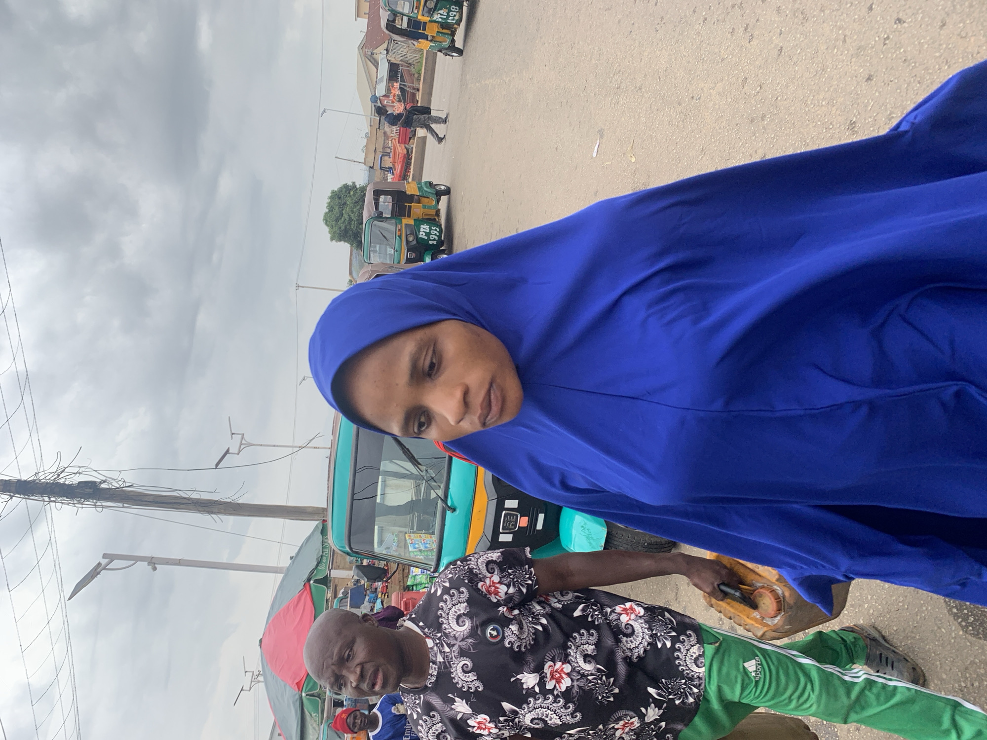 The image shows a woman wearing a blue hijab, partially obstructing the view, and a man in a black and white floral shirt and green Adidas track pants. The man carries a yellow jerrycan. In the background, there are several green and yellow auto rickshaws (tricycles), overhead power lines, and other indistinct people and market stalls, suggesting a busy street scene in Jos, Nigeria. The sky is overcast.