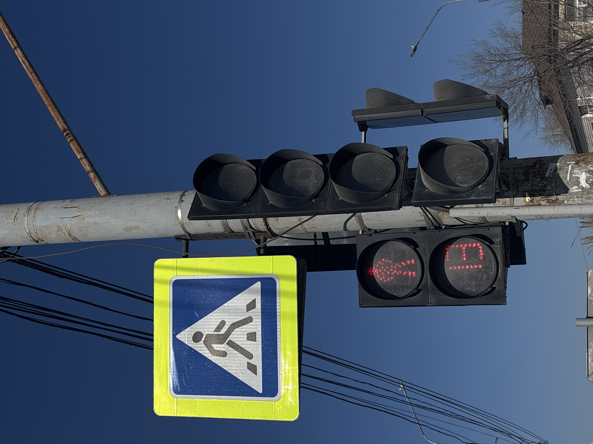A low-angle shot shows a pedestrian crossing signal and traffic lights against a blue sky in Perm, Russia. The pedestrian sign features a blue square with a yellow border and a white triangle containing a black figure walking across white bars. To the right, a pedestrian traffic light displays the red countdown "19." Above the pedestrian light are four unlit traffic lights, with an additional two above them. The structure supporting the lights and sign is gray and shows signs of wear. Power lines crisscross the lower portion of the frame. A building with trees is visible in the upper-right corner.