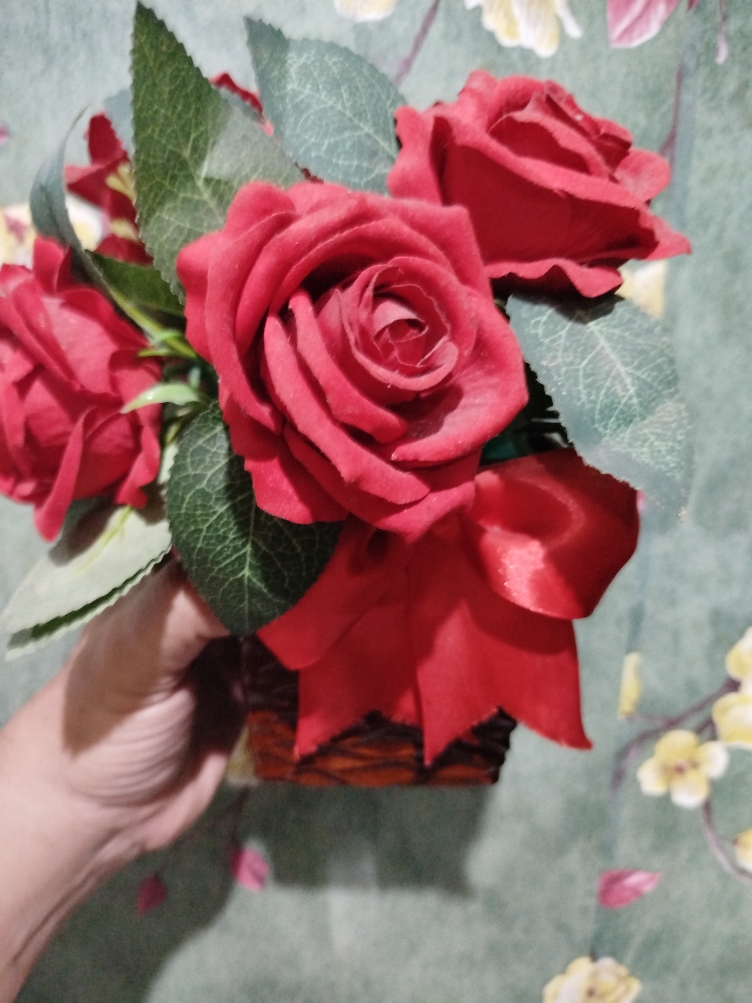 A hand holds a small, woven basket containing a bouquet of artificial red roses and green leaves. A red satin ribbon is tied around the basket. The background is a muted green wall with a floral pattern of yellow and pink flowers. The scene appears to be indoors, in a residential setting.