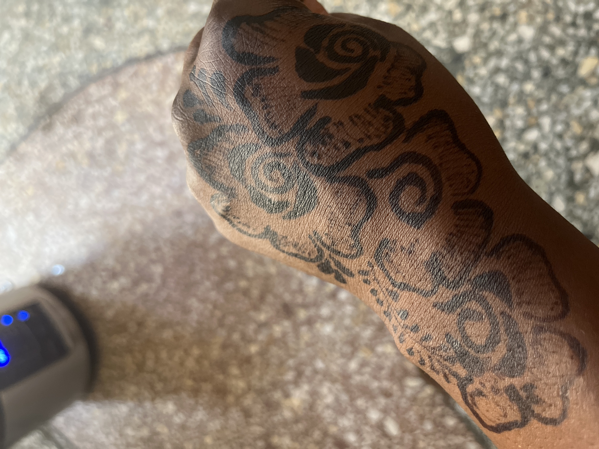 A close-up shot shows a dark-skinned human hand with a dark henna-like design covering the top. The design appears to be a series of floral patterns and swirls. The background shows a blurred surface, possibly concrete or a tiled floor, and a portion of an electronic device with blue lights. There is no way to tell the location.
