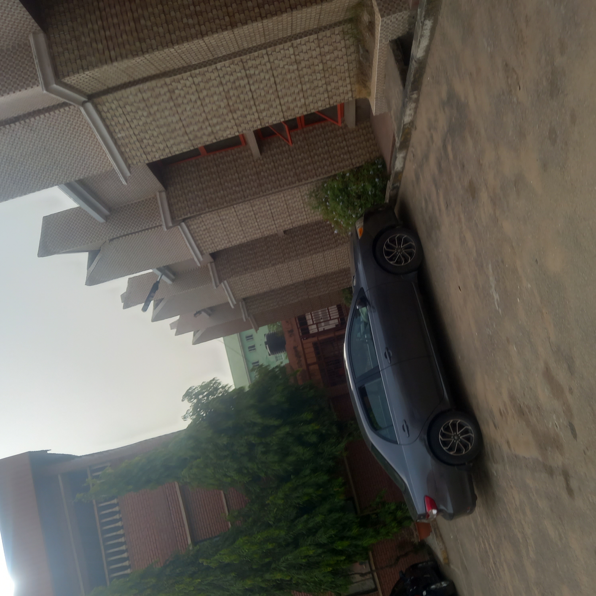 The image displays an outdoor scene captured during the daytime, rotated 90 degrees clockwise.
The central subject is a dark grey sedan, parked on a light brown or greyish-brown paved surface, possibly a road or parking lot. The car faces towards the right of the frame (initially the top of the image), featuring multi-spoke alloy wheels.
In the background, on the left side of the rotated image (originally the top), a multi-story building with a distinctive textured facade is prominent. This building features rows of rectangular, light brown or beige tiles arranged in a checkerboard or woven pattern. Projecting architectural elements, possibly sunshades or decorative eaves, extend outwards from the building's upper levels, creating a stepped appearance. Orange window frames are visible on some parts of this building.
Further in the background, partially obscured by lush green trees and bushes, is another building painted in a light green color with white windows. A reddish-brown building with vertical paneling is also visible on the lower left (originally the bottom), surrounded by dense green foliage.
The sky, visible in the upper left (originally the bottom), appears bright and overcast. No human activity or specific event is depicted. There is no visible text.