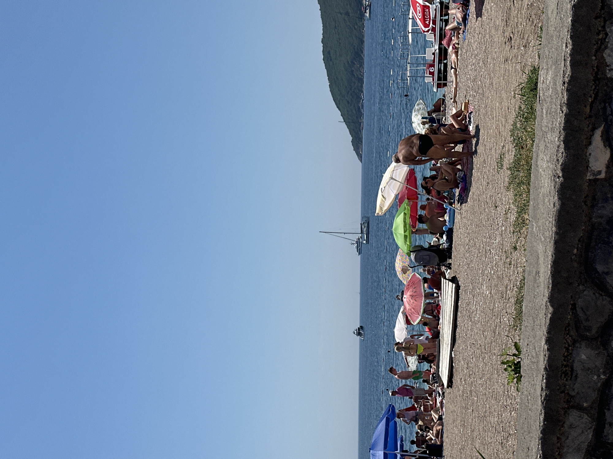 The image depicts a crowded beach scene on a sunny day. The main subjects are numerous people relaxing and sunbathing on a pebbly shore. Objects visible include a variety of colorful beach umbrellas, beach chairs, and a few boats or yachts in the blue sea. The scene is set against a backdrop of a green, tree-covered hillside that slopes down towards the water. The bright blue sky suggests clear weather and likely midday, given the strong sunlight casting shadows. The presence of numerous people indicates a popular tourist destination, and the overall atmosphere appears relaxed and leisurely, consistent with a beach vacation. The location is identified as Budva, Montenegro. There is some visible text on a red and white umbrella, which appears to read "ALGIDA".