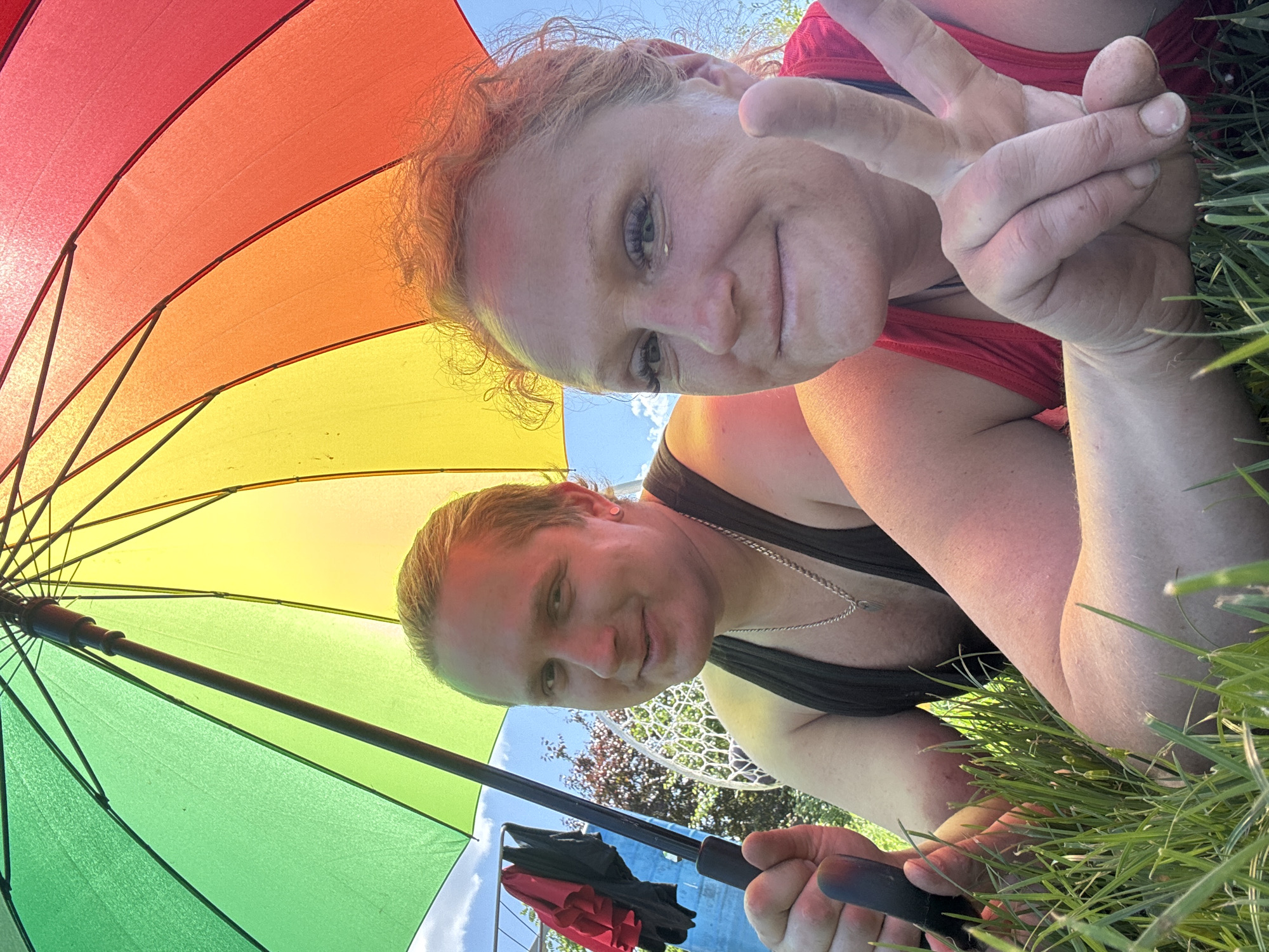 Two individuals are lying in the grass under a rainbow-colored umbrella. The person on the left, with blonde hair pulled back, wears a black tank top and a silver chain necklace. They are holding the umbrella handle. The person on the right, with reddish, curly hair, wears a red tank top and is making a peace sign with their right hand. Both individuals are smiling at the camera. In the background, there is a clothesline with garments, trees, and a white outdoor chair. The scene appears to be a backyard or park setting. The location in Chaikovsky, Russia is not discernible from the image.