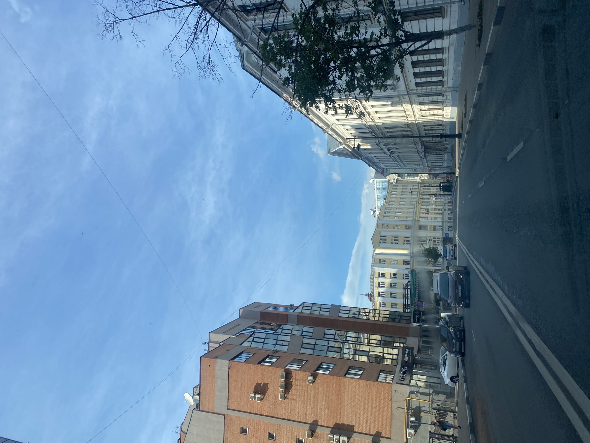 The image captures a street scene in Novokuybyshevsk, Russia, under a bright blue sky with wispy clouds. The perspective is from a low angle, looking upwards. On the left side of the street, two multi-story buildings dominate the frame. The closer building is a modern brick structure with numerous windows and several outdoor air conditioning units attached to its facade. Further down the street, another, older building with a white facade and many windows is visible, partially obscured by a tree with bare branches.
On the right side of the street, a paved road runs alongside the buildings, marked with white lines. Several cars are parked and driving on the road, including a dark-colored SUV and a white sedan. A few pedestrians are visible on the sidewalk, one walking and another near a fence. The overall impression is a typical urban environment on a clear day, possibly in the late morning or early afternoon, given the quality of the sunlight. The presence of air conditioning units suggests it might be a warmer season. No specific emotional cues are evident in the static image. A sign on the brick building on the left reads "KRIM KITCHEN" and another smaller text below it reads "illa-design".