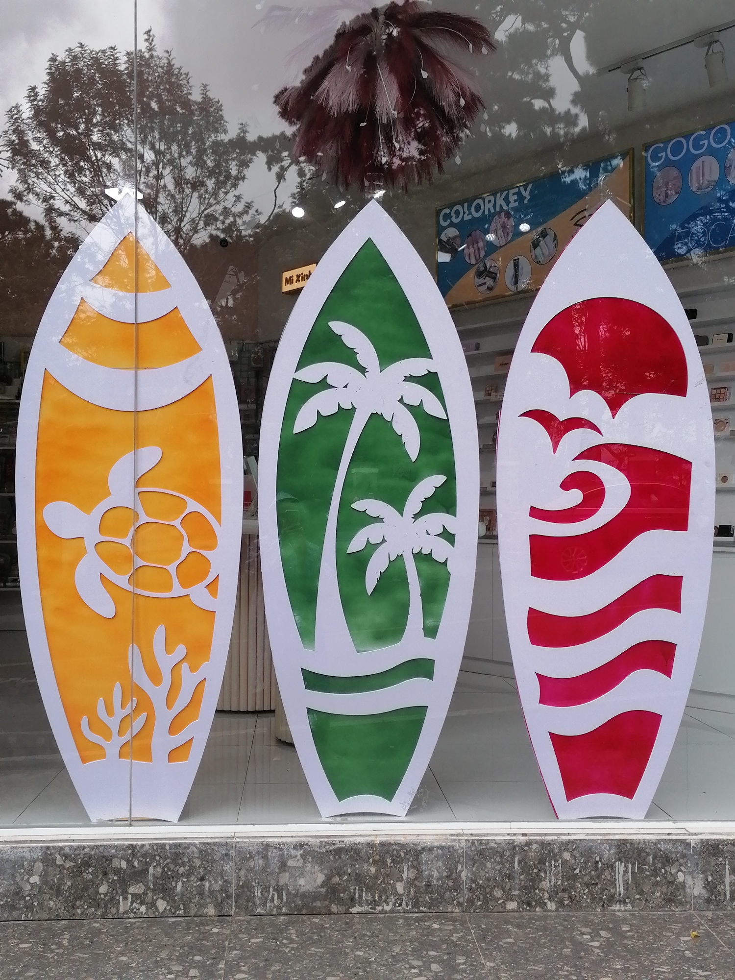 In Da Lat, Vietnam, three surfboard-shaped decorative panels are displayed in a shop window. The leftmost panel is orange with a white silhouette of a sea turtle and coral. The middle panel is green with white silhouettes of palm trees. The rightmost panel is red with white wavy lines and two bird silhouettes. The panels are positioned side-by-side against a reflective glass surface, showing reflections of trees, a feathered decoration, and posters on the shop interior. The background inside the shop appears to be a retail setting. The scene is viewed from a sidewalk with a textured grey stone base.