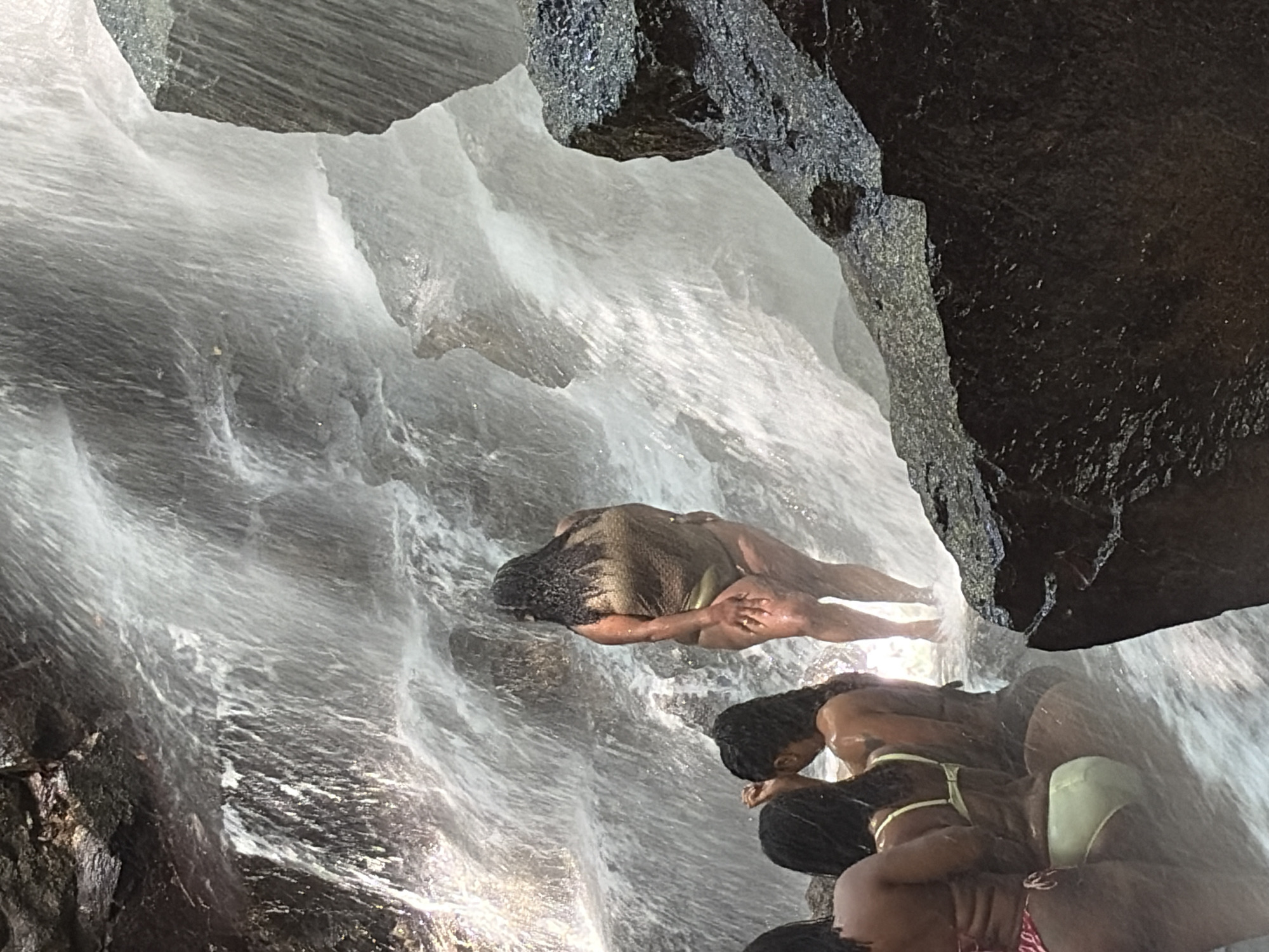 The image depicts a group of individuals at a waterfall. In the foreground, on the left side, three people are partially visible, wearing bikinis. The person furthest to the left is wearing a pink and white bikini. The person next to them is wearing a yellow bikini. The person on the far right of this group is also in a bikini, with dark hair. In the center of the image, a person in a gold, textured bikini stands facing away from the camera, looking towards the waterfall. The background is dominated by the cascading water of the waterfall, with rocks visible among the spray. The environment appears to be natural and rocky, with lush greenery suggested by the darker areas of the rocks. The water is the primary element of the environment, creating a dynamic and refreshing atmosphere. The location is identified as a waterfall in Gianyar, Indonesia.