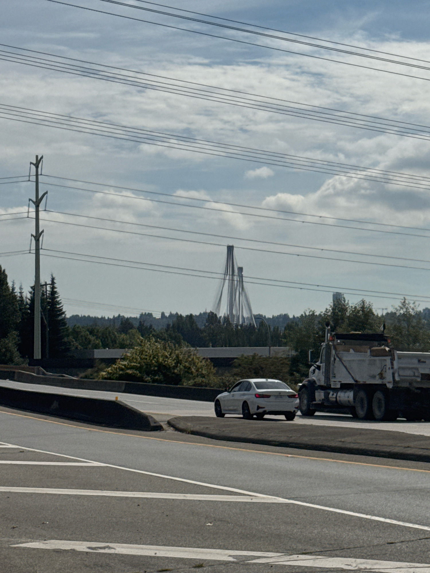 This image depicts a view of the Alex Fraser Bridge in Coquitlam, Canada, under a cloudy sky. In the foreground, a white BMW sedan is driving on a highway. To its right, a white dump truck is also in motion. The middle ground features a dense line of trees. In the background, the distinctive, splayed cable-stayed towers of the Alex Fraser Bridge rise above the tree line, with a single high-rise building visible to the right of the bridge. Overhead power lines crisscross the sky, supported by a tall utility pole on the left side of the frame. The overall scene suggests an urban or suburban setting with significant infrastructure.