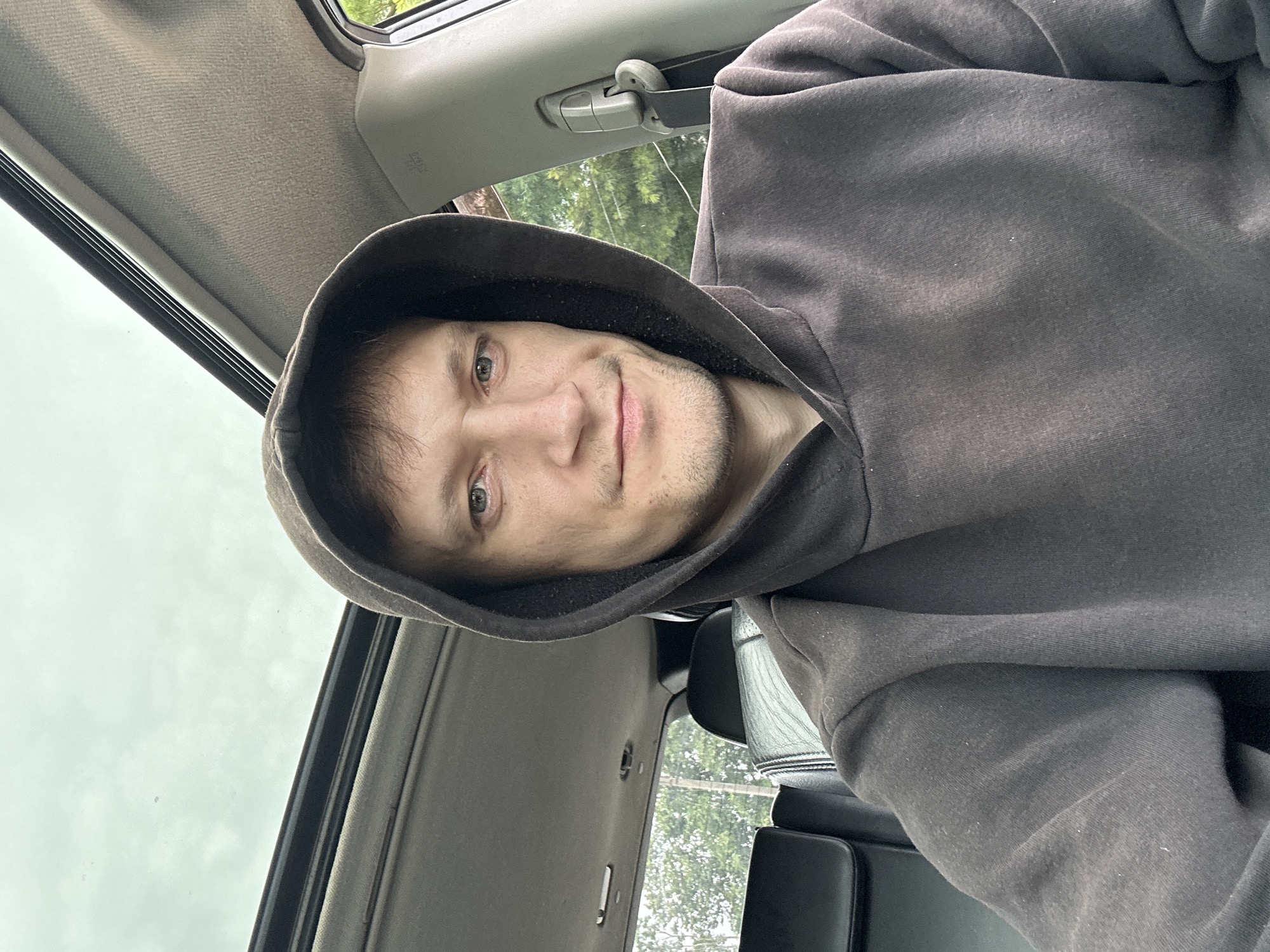 A man wearing a dark hooded sweatshirt is seated in the driver's seat of a car, looking towards the camera. The car's sunroof is open, revealing a cloudy sky. The interior of the car is visible, including the headliner and a portion of the passenger seat. Outside the car, trees and foliage can be seen. The man's face is clearly visible, and he has a faint smile. There is no indication that this scene is located in Makiv, Ukraine.