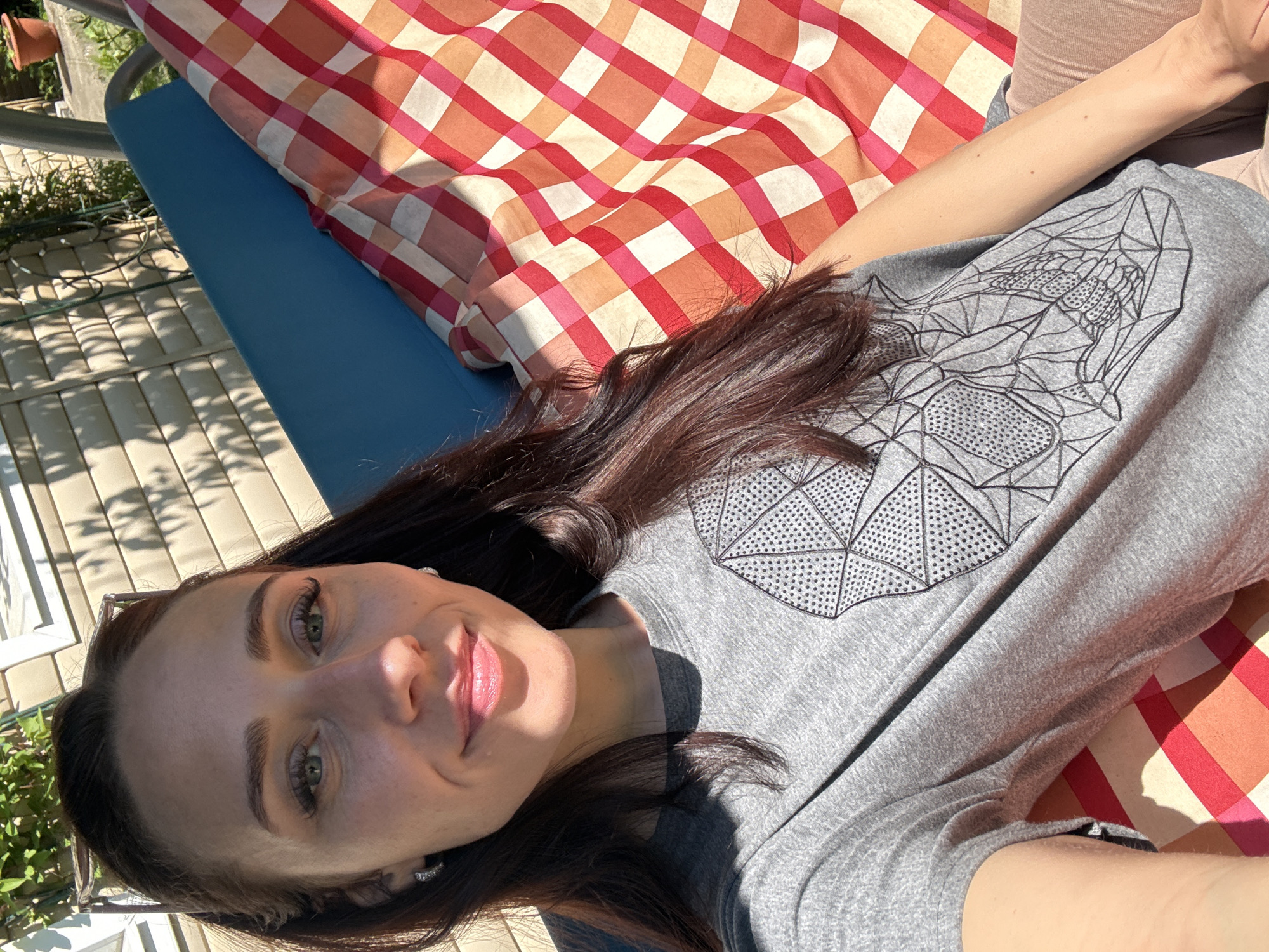 An adult female, approximately in her 20s or 30s, is captured in a selfie outdoors in the city of Saint Petersburg, Russia. She has dark brown hair, pulled back with sunglasses perched on her head, and is wearing small, silver-toned hoop earrings. Her eyes are green, and she presents a slight smile with visible lip gloss. She is clad in a grey, short-sleeved t-shirt featuring a black geometric skull design outlined and partially filled with small, dark, studded embellishments. Her lower attire consists of light-colored, possibly tan or light pink, pants.
The woman is seated on a blue-framed garden swing, which is partially covered by a red, white, and orange plaid fabric serving as a cushion or blanket. The scene is illuminated by strong sunlight, evidenced by distinct shadows cast across the woman's face, shirt, and the background elements. The background features light-colored, horizontal wall siding from a building, with green foliage visible above and to the right. A terra cotta-colored potted plant is discernible in the upper right background, and a paved or concrete surface is partially visible beneath the swing.