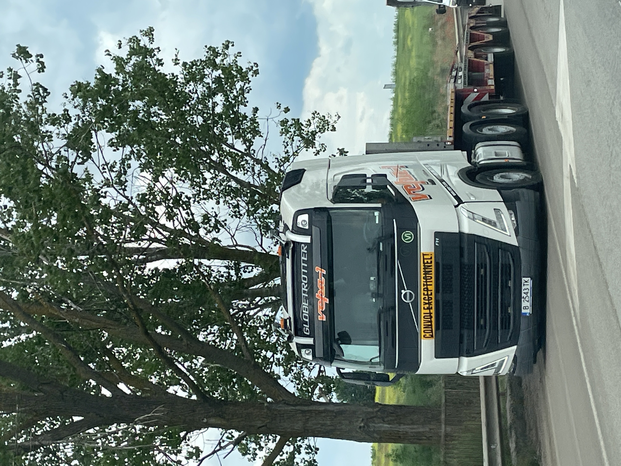 A white Volvo FH truck with a Romanian license plate B 2543 TK is parked on a road. A large tree is to the left of the truck. A flatbed trailer is attached to the truck. The surroundings suggest a rural or roadside location, potentially near Kamenar, Bulgaria. The truck features "GLOBETROTTER" branding and signage indicating "CONVOI EXCEPTIONNEL!" There are no visible people in the image. The sky is partly cloudy.