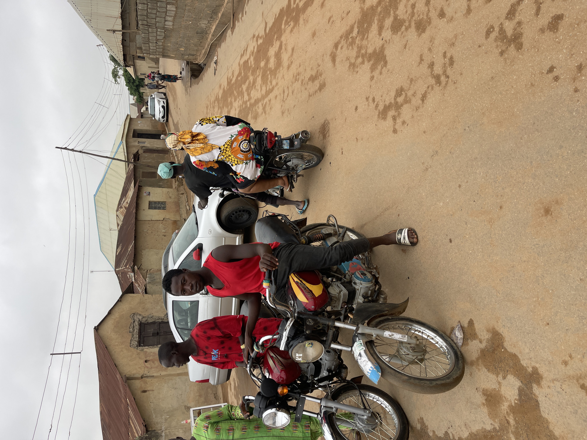 Two men are seated on motorcycles on a dirt road in what appears to be Kuchingoro, Nigeria. The road is flanked by buildings of varying construction, and a white car is parked behind the motorcycles. One man is wearing a red tank top and black pants, and the other a red patterned shirt. A third motorcycle is partially visible in the foreground. Other people and a car are seen farther down the road. The sky is overcast.