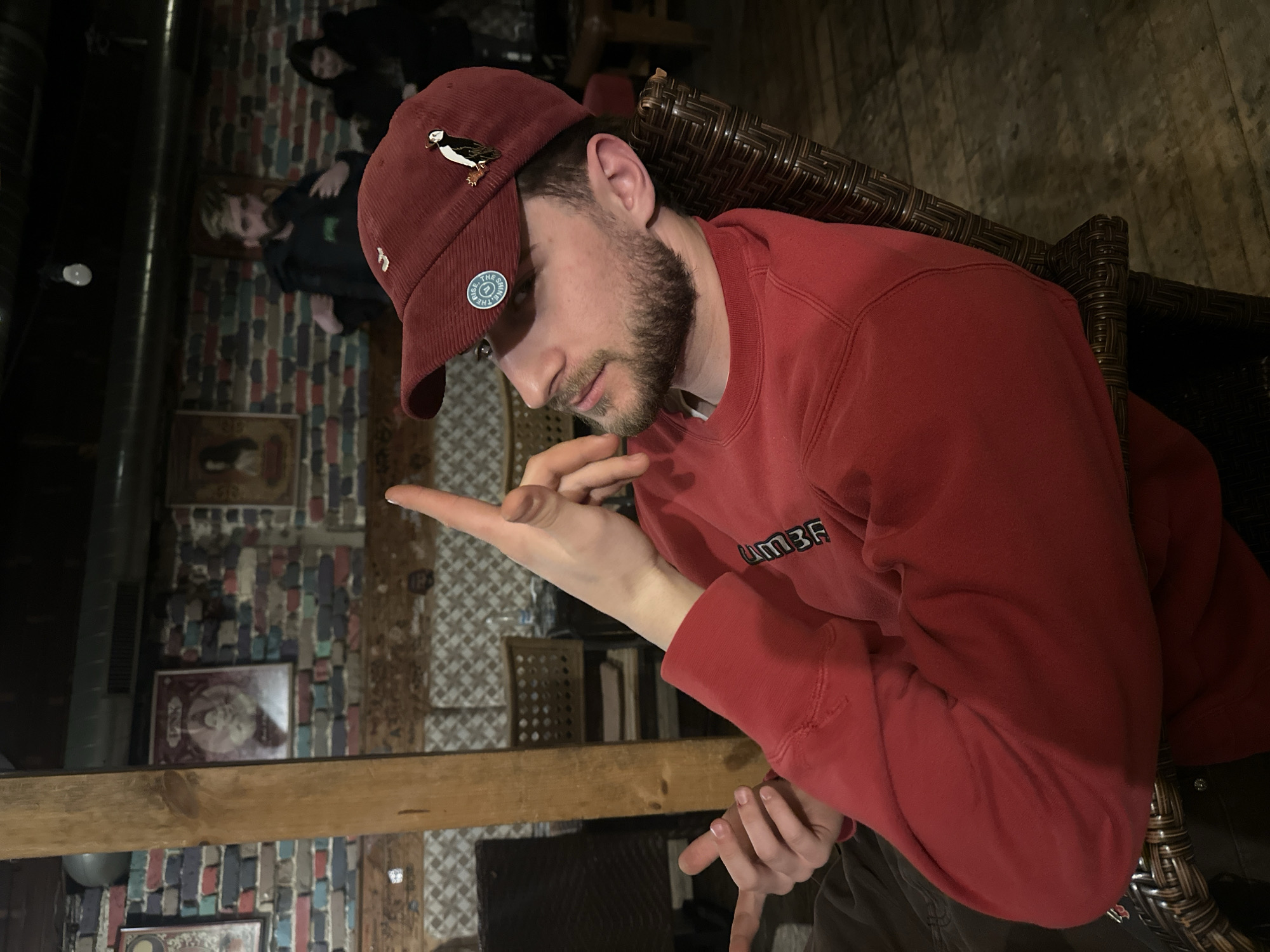 A male individual with short brown hair and a beard, wearing a red corduroy baseball cap and a red sweatshirt, is seated in a dark brown woven wicker-style chair. His left arm is raised with the index finger extended upwards. His head is tilted slightly back and to the left, with a neutral expression. The baseball cap features a circular blue and white pin with "SHINE ON & RISE UP" text and an enamel puffin pin. The sweatshirt has "UMBRA" text visible on the chest.
The scene is an interior location in Plovdiv, Bulgaria, characterized by a distressed multi-colored brick wall in the background, adorned with framed artwork. A prominent vertical wooden beam is positioned to the left of the main subject. Overhead, a metal ventilation duct is visible, and lighting consists of exposed bulbs. Another male individual with a beard, wearing a dark jacket, stands in the midground, appearing to adjust his clothing. A female individual with dark hair is partially visible further in the background to the right. Various dark brown wicker chairs and tables are present throughout the dimly lit space. A clear glass bottle stands on a table in the background. The floor is dark wood.
