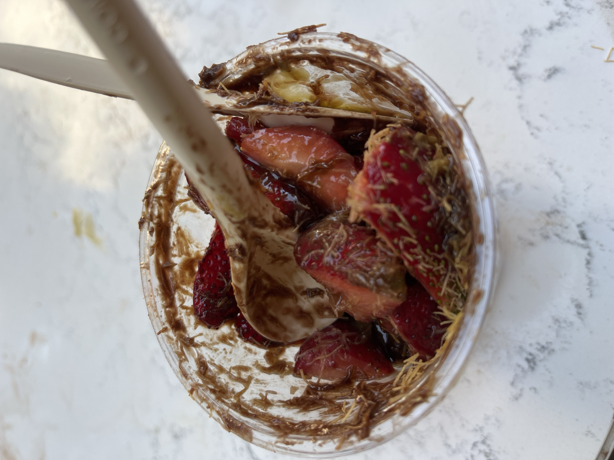 A top-down view shows a clear plastic cup filled with strawberries, banana slices, and what appears to be chocolate sauce or paste. A light-colored, disposable spoon rests inside the cup, partially obscured by the contents. Shredded noodles or similar textured food items are also mixed in. The cup sits on a white surface with a marbled pattern. The location is unspecified and therefore cannot be identified.