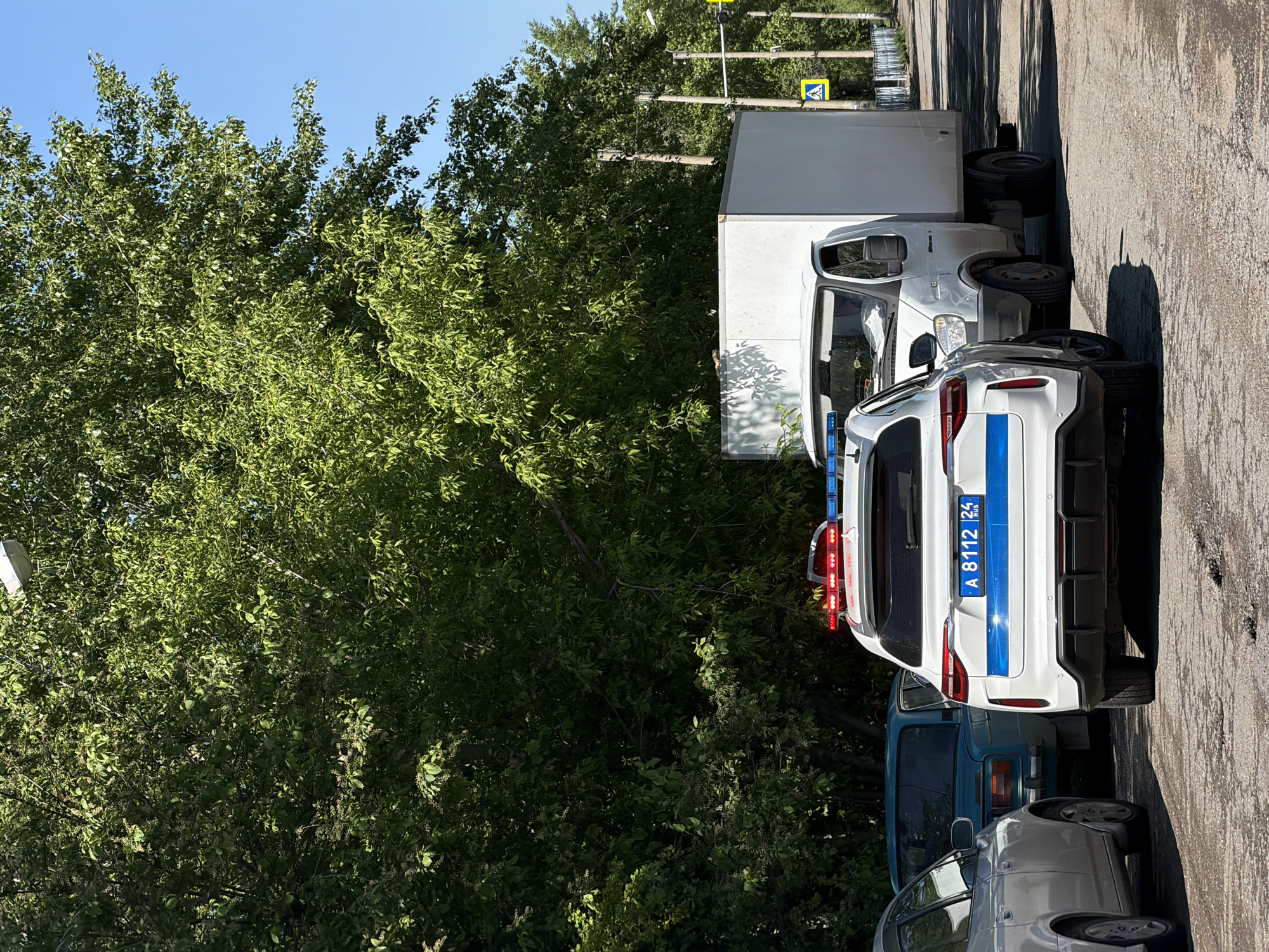 A parked white Russian police car, license plate "A 8112 24 RUS", is seen from the rear on a city street in Beryozovka, Russia. The car is flanked by a white box truck to the right and two other vehicles, one dark blue and one silver, to the left. Lush green trees fill the background, partially obscuring a street sign. The location is a street-side parking area with an asphalt surface. The time of day is daytime, with bright sunlight creating shadows.