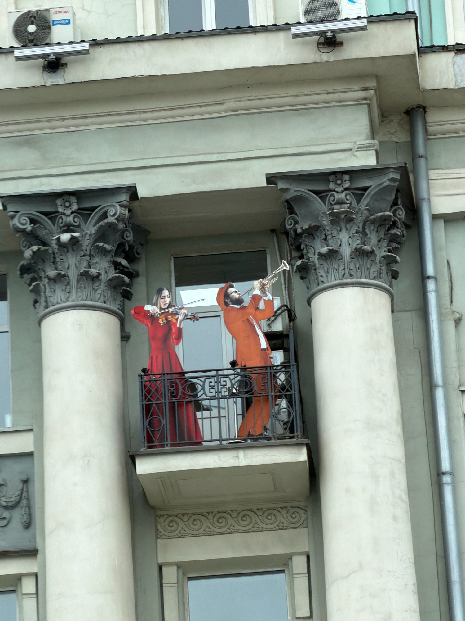 The image shows a balcony on a building in Moscow, Russia. Two cardboard cutouts of people are standing on the balcony. One cutout is of a woman in a red dress playing a violin. The other cutout is of a man in an orange suit playing a trumpet. The building is light green with decorative columns. An air conditioning unit is visible on the roof of the building.