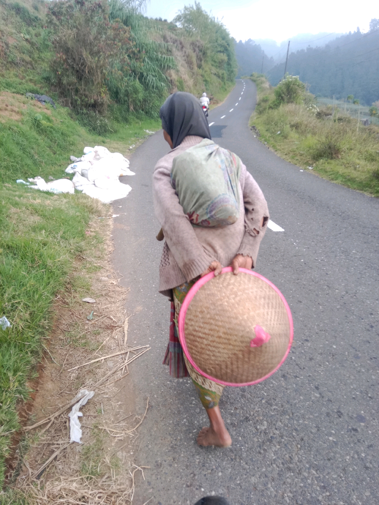 A woman, wearing a dark hijab, a light brown sweater, and a patterned skirt, walks along a paved road. She carries a conical straw hat with a pink rim and a pink accent on top. A baby is wrapped in a light green cloth and carried on her back. The road winds through a hilly, rural landscape with green vegetation and trees. In the background, a motorcycle is visible further up the road. To the left of the road, there is a grassy embankment with several white sacks piled up and some scattered debris. The scene appears to be set in Batur, Indonesia, given the distinctive conical hats and the mountainous terrain characteristic of the region.