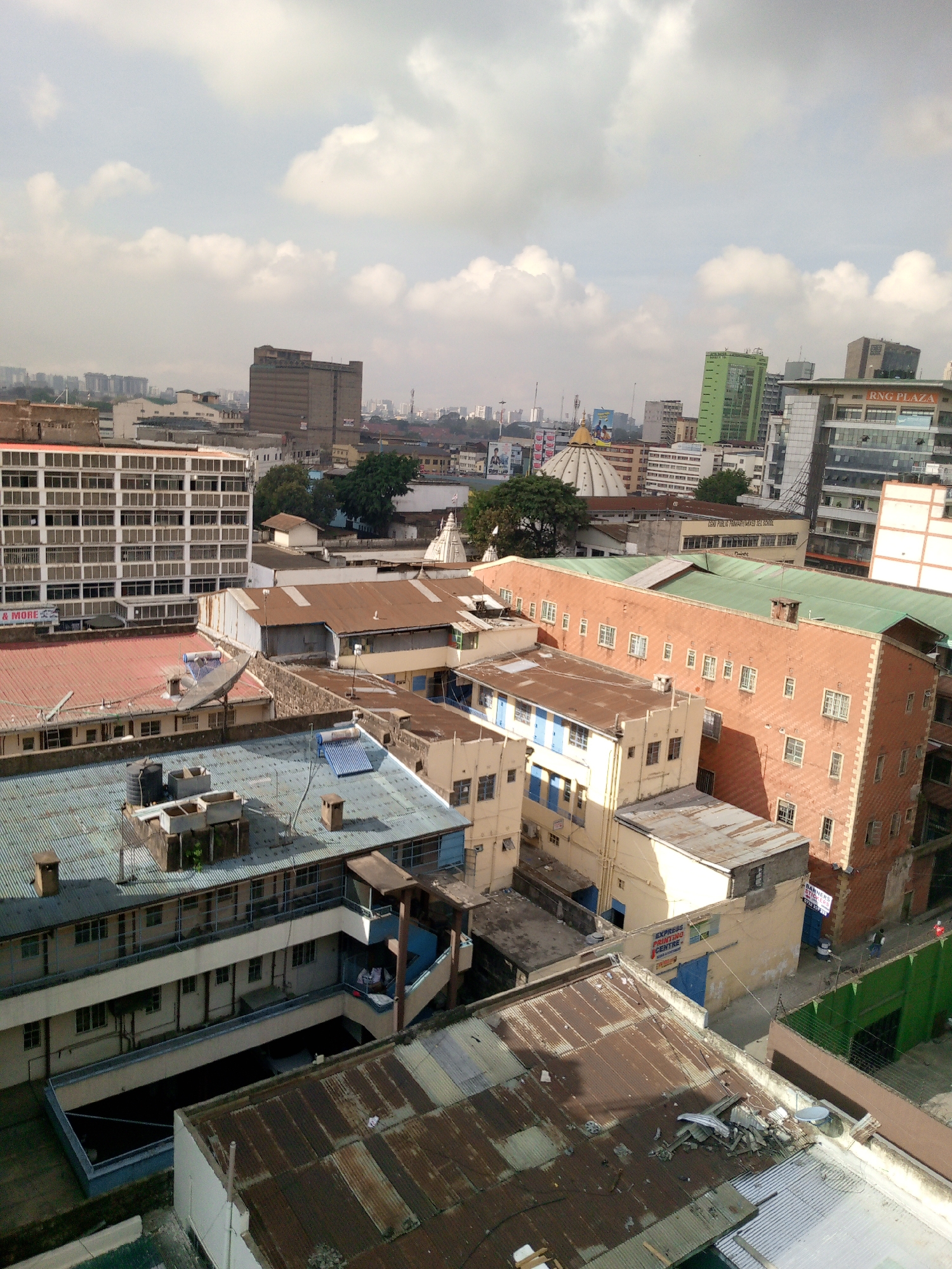 The image depicts a high-angle view of a dense urban landscape in Nairobi, Kenya, under a partly cloudy sky. The foreground features a collection of older buildings with corrugated metal roofs, some showing signs of rust and age. Various structures including a building with a courtyard. Moving into the midground, are taller buildings with a combination of red, green, and metallic roofs, and there is a Hindu temple that is partially visible. The background shows modern high-rise buildings, including a green one. The overall scene conveys a mix of old and new architectural styles.