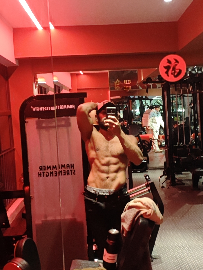 A gym interior in the Chongqing High-tech Zone, China, features red ambient lighting and red-painted walls. A shirtless, bearded male, wearing a black cap and dark pants, is centered, taking a mirror selfie. He possesses a muscular physique with visible abdominal definition and a piercing on his left nipple. His left hand, holding a smartphone, partially obscures his face. A 'HAMMER STRENGTH' branded weight machine (text reversed in mirror) stands to the subject's left. Reflected in the background mirror, another individual in a light-colored jacket and pants is partially visible amidst black-framed gym equipment. A red circular sign with the black Chinese character '福' (fú) is mounted on gym equipment in the upper right. A black bottle with a red label and a light-colored towel are positioned on gym equipment in the foreground, right of the subject. The floor is textured.