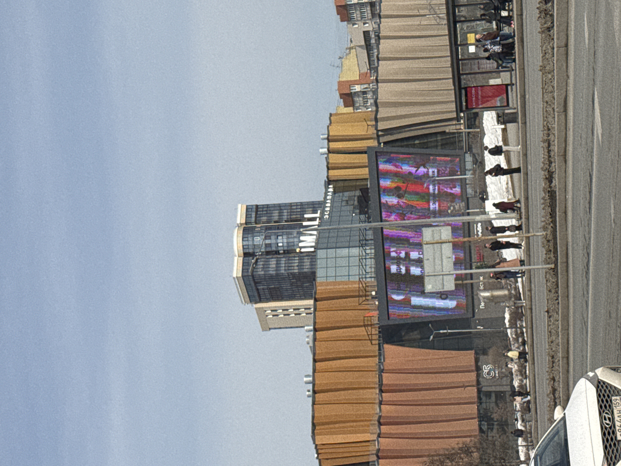 The image depicts a daytime scene in Perm, Russia, specifically on Shosse Kosmonavtov (Highway Cosmonauts), featuring the Planeta (iMALL / Escalada) shopping and entertainment complex.
The central subject is a large, modern commercial building with distinctive architectural elements. The main mall structure features a facade composed of numerous vertical, corrugated, or folded panels in varying shades of orange and brown. Above and behind this structure, a taller, multi-story building is visible, constructed primarily of glass and dark-toned panels, bearing prominent signage for "iMALL" and "ЭСКАЛАДА" (ESCALADA). A large, vibrant LED screen on the mall's lower facade displays abstract, colorful digital imagery.
In the foreground, a multi-lane asphalt road is present, separated by a concrete median strip. A white Hyundai sedan, partially visible on the left, occupies the nearest lane, displaying Russian license plate "P864AH 159", indicating the Perm Krai region. To the right of the road, a bus stop with a glass shelter is occupied by approximately ten individuals. Several other pedestrians are distributed along the sidewalk and a snow-covered embankment between the road and the mall's entrance. These individuals are dressed in winter or early spring outerwear, including dark jackets and coats. Patches of snow and bare trees are visible, indicating cold weather conditions, likely late winter or early spring. The sky is clear and light blue. Streetlights, traffic signs, and trash receptacles are positioned along the pedestrian areas.