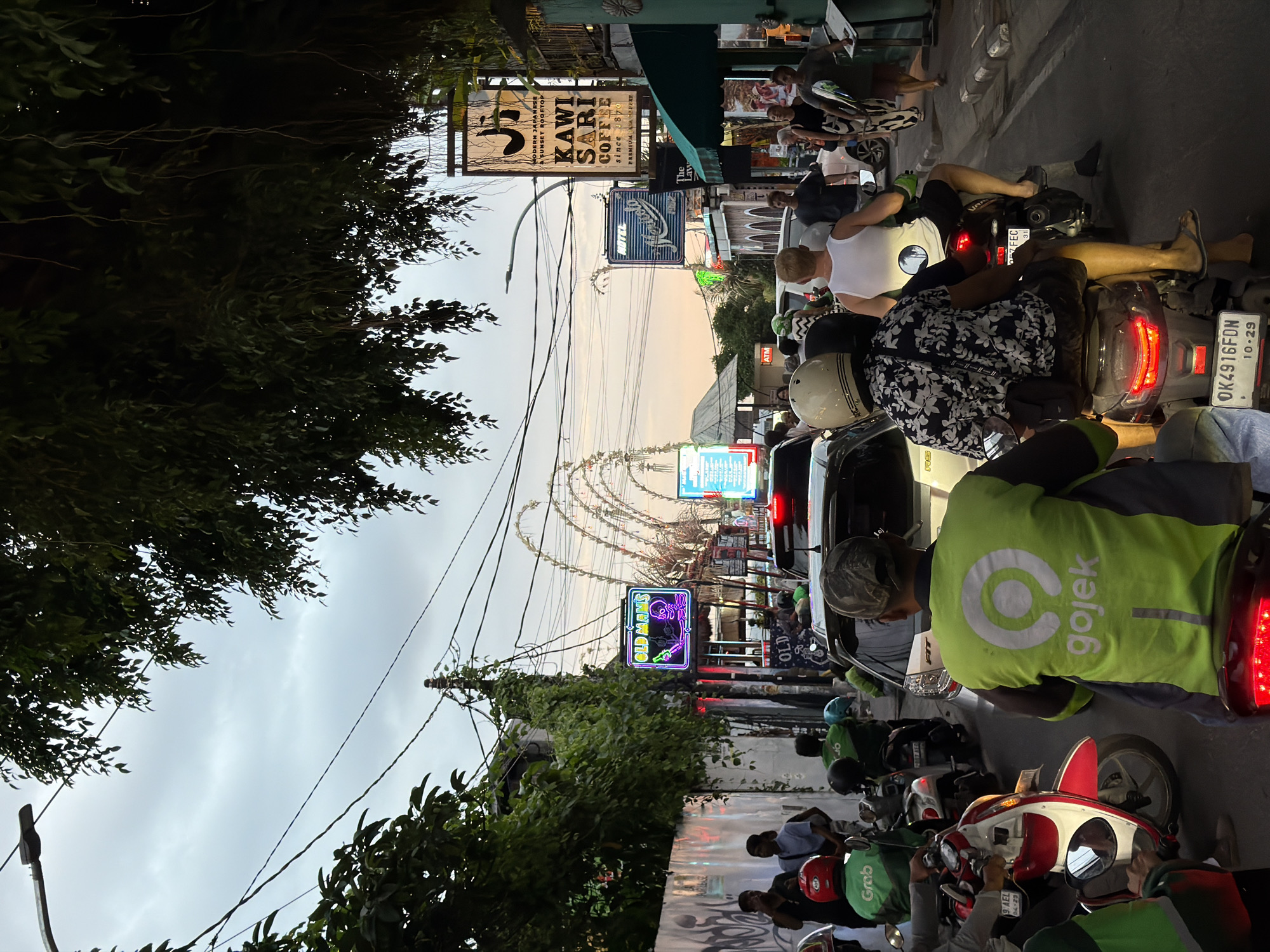 The image depicts a congested street scene in North Kuta, Indonesia, during what appears to be dusk. The road is filled with motorbikes and cars, indicating heavy traffic. Several individuals are on motorbikes, many wearing helmets. Some are clad in bright green vests with the "Gojek" logo, suggesting they are delivery or ride-sharing drivers.
On the right side of the street, businesses are visible, including one with a prominent sign that reads "KAWI SARI COFFEE since 1870." Another sign for a "HOTEL" is also present. Elaborate, decorative archways, possibly part of a local festival or decoration, are visible in the background, extending over the street. Trees with dense foliage line the sides of the road, casting shadows and contributing to the atmospheric lighting, which is further enhanced by neon signs. The sky is overcast, with a subtle gradient indicating the transition from daylight to evening. The overall impression is a lively, bustling urban environment characteristic of a popular area in Indonesia.