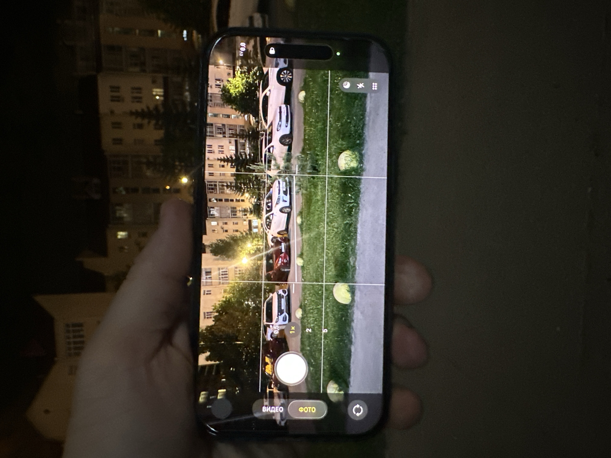 The image depicts a person holding a smartphone, presumably taking a photograph. The smartphone screen displays the camera app in photo mode, with the viewfinder showing a scene at night. The scene includes a residential building with lit windows, several parked cars, a grassy area, and a paved sidewalk. The camera app displays gridlines, indicating a potential photographic composition aid. The phone model is an iPhone based on the UI. The image was likely taken in Kazan, Russia, based on the text displayed on the screen.