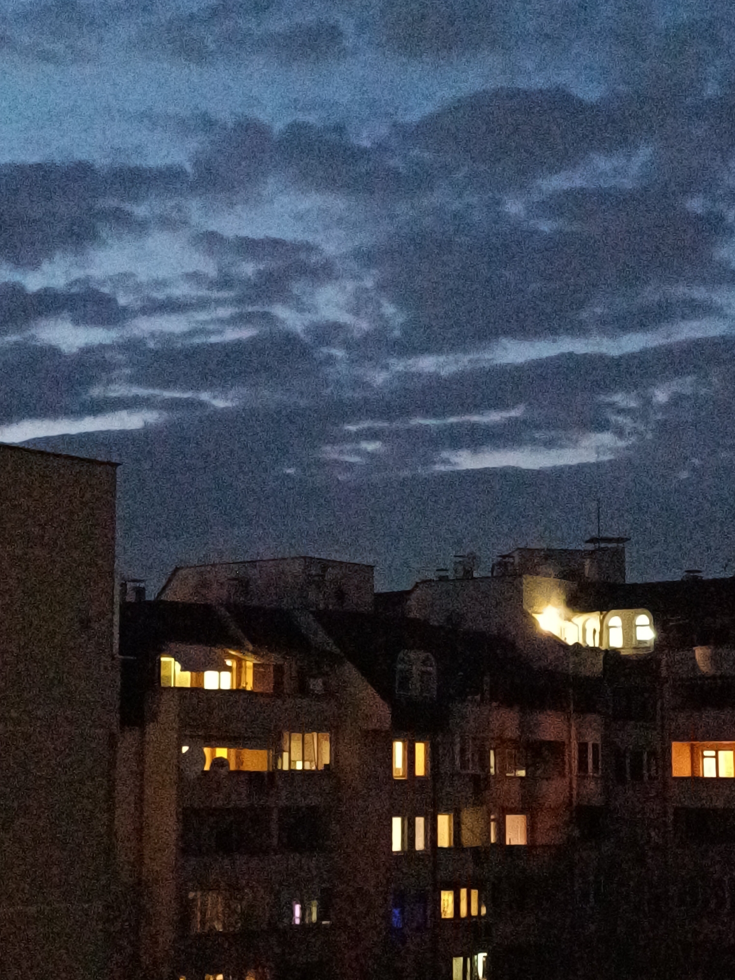 The image presents an urban area in Plovdiv, Bulgaria, during evening or night hours. A dark, heavily clouded, blue-grey sky occupies the upper frame. The lower two-thirds display a dense arrangement of multi-story buildings. A tall, unlit, concrete-textured building is partially visible along the left edge. The central and right sections reveal a complex of buildings with diverse roof structures, including chimneys and a prominent rooftop antenna on one building. Multiple windows across these structures are illuminated with warm yellow light. A building in the upper-center-right features several brightly illuminated arched windows. No human figures are discernible. The image exhibits significant visual noise, consistent with low-light capture.