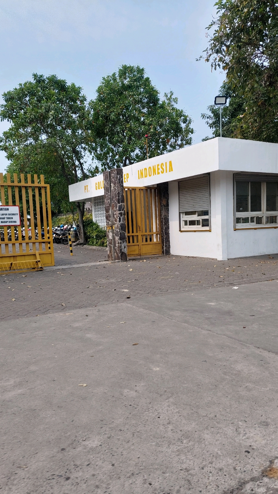 The image depicts the entrance to PT. Golden Indonesia in Pilang, Indonesia. The entrance features a white building with the company name in yellow text above the entrance. A yellow metal gate is partially open, revealing a parking lot with motorcycles. The ground is paved with concrete and cobblestones. Trees surround the entrance, and the sky is visible above. A sign is posted on the yellow gate with text in Indonesian. The scene is well-lit, suggesting it was taken during the daytime.
