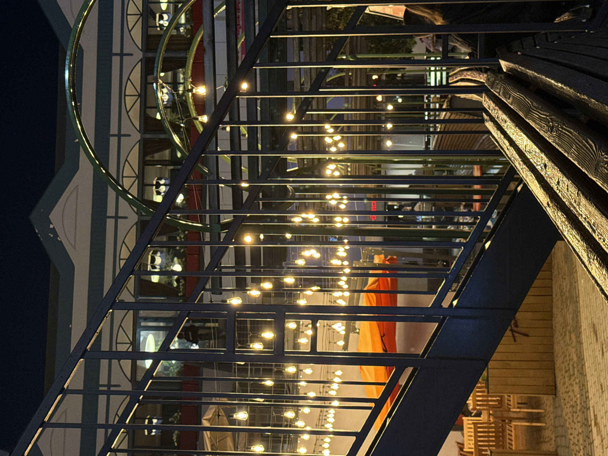 The image depicts an outdoor area at night, likely a public space like a cafe patio or a park. A dark blue metal staircase with railings is prominently featured in the foreground, leading upwards and to the right. Below the staircase, there is a wooden partition with what appears to be graffiti on it, and some wooden chairs and tables are visible further back.
The background reveals a building with arched windows and a light-colored facade, adorned with green and white striped trim. Numerous small, warm-toned lights are strung across the scene, illuminating various sections. These lights are either hanging individually or integrated into decorative green arches. The overall ambiance is warm and inviting, suggesting a place open for evening activity. While the precise location is not definitively identifiable from the image alone, the prompt specifies Ufa, Russia, hinting at a potential urban setting within that city. No specific actions or interactions between people are clearly visible, though indistinct figures can be seen in the background.