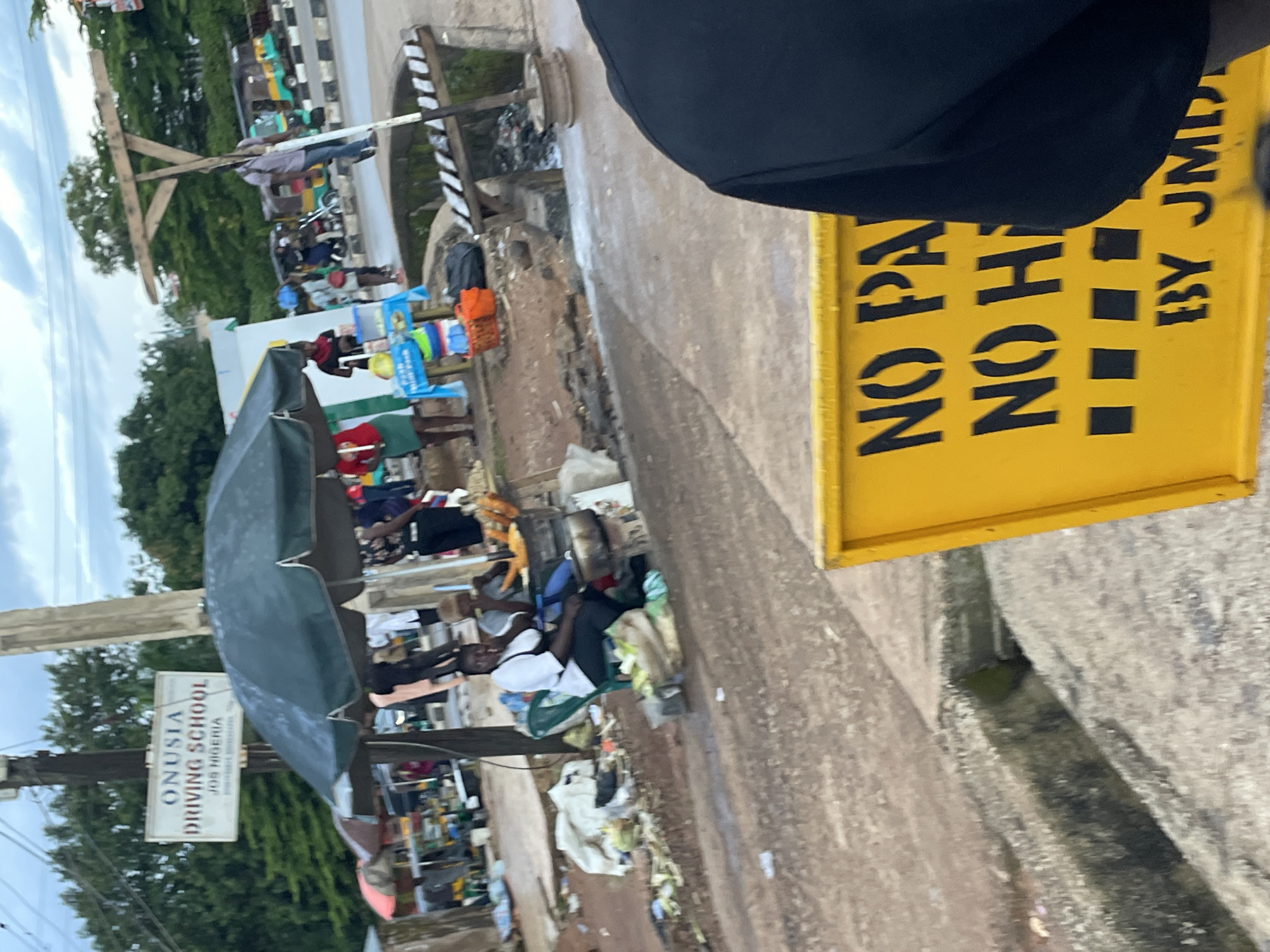 This image captures a street scene in Jos, Nigeria, featuring a bustling roadside with vendors and pedestrians. In the foreground, a bright yellow sign with black stenciled lettering reads "NO PA NO H BY JMD," indicating a restriction on parking or some other activity, with the final part of the text obscured. Behind the sign, a drainage channel runs along the road, with some debris and water present.
Further into the scene, a driving school sign is prominently displayed, reading "ONUSIA DRIVING SCHOOL JOS NIGERIA," suggesting a nearby driving academy. Several vendors are set up under a large, dark umbrella, selling various goods. People are visible engaging in activities such as sitting, standing, and presumably conducting business. In the background, three-wheeled vehicles, common in Nigeria, are seen on the road, along with more trees and buildings, indicating a typical urban environment. The overall atmosphere appears to be one of everyday commerce and transit in Jos.