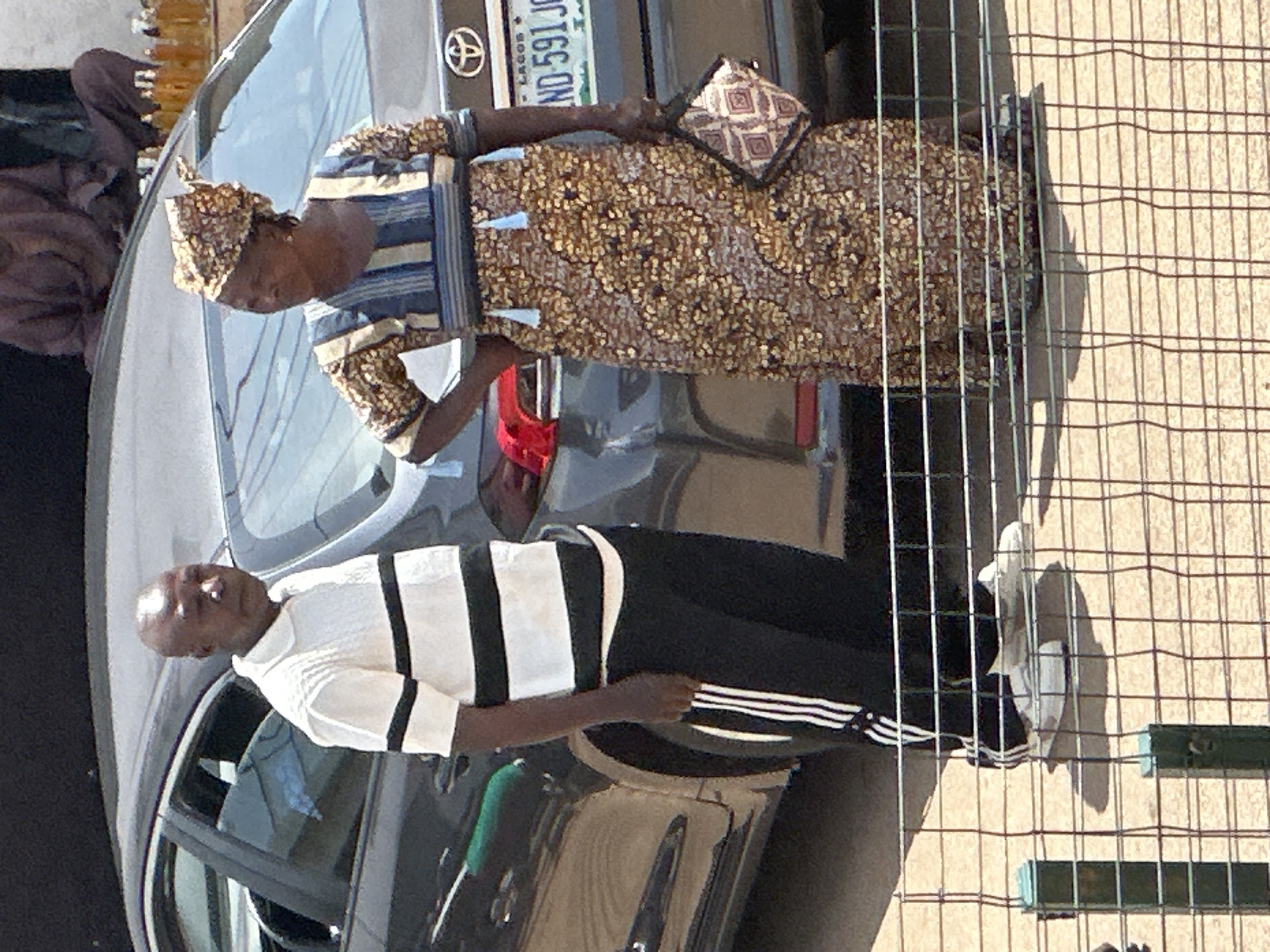 In Bukuru, Nigeria, a man and a woman stand next to a parked car on a paved surface, possibly a roadside. The man, with a bald head and dark skin, wears a white polo shirt with horizontal black stripes and black athletic pants with white stripes. He is looking down. The woman, also with dark skin, wears a patterned dress and a headwrap. She has her hand on her hip and is looking to her left. A patterned clutch bag is held in her right hand. The car appears to be a dark gray sedan, with a license plate visible that reads "LAGOS ND 591 JC". In the background, a person in a brown robe is seated behind the car, and rows of small bottles with amber liquid are displayed. A wire fence with green posts runs across the foreground of the image.