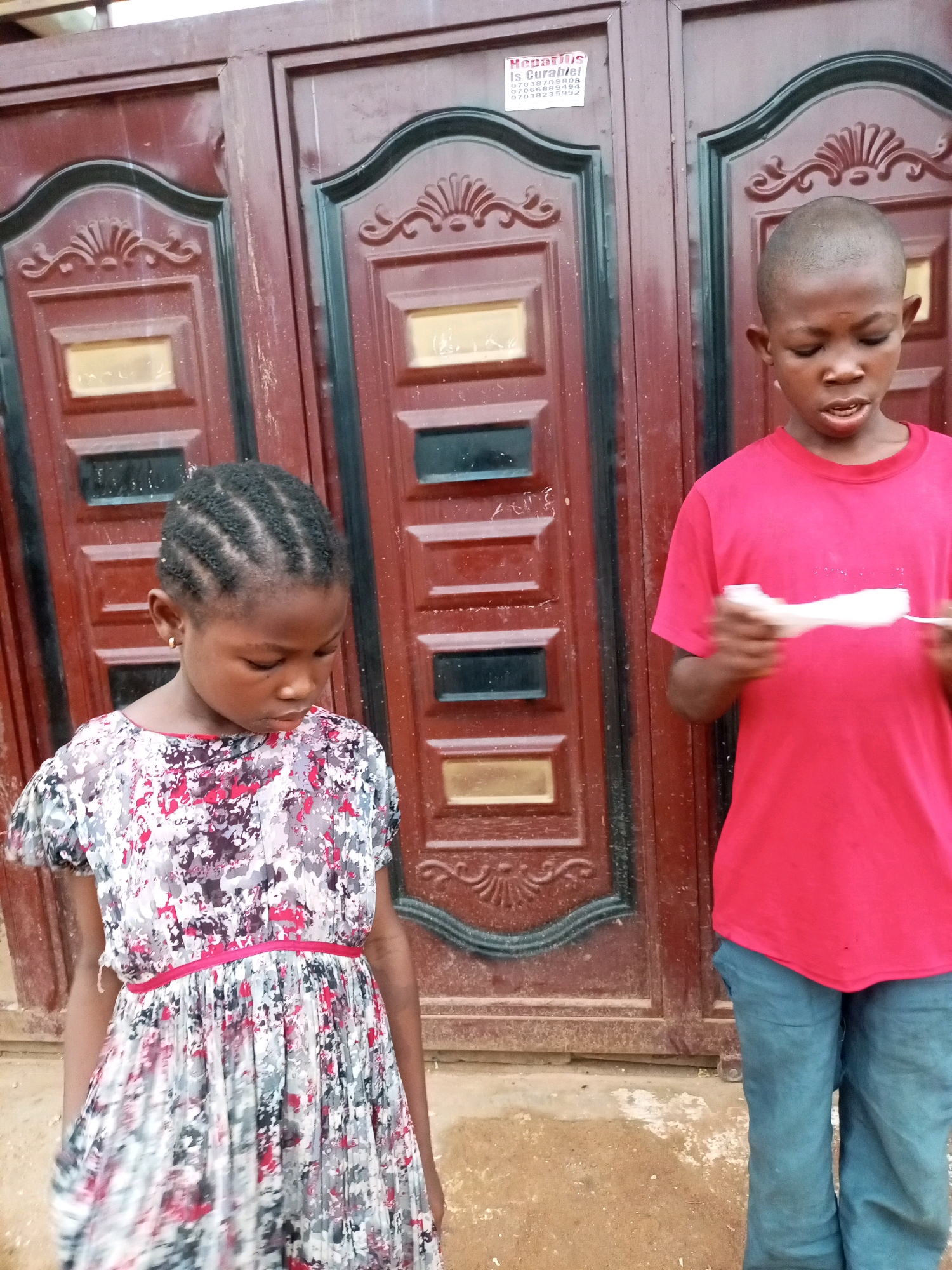 In Tempure, Nigeria, two young individuals stand in front of a large, ornate, dark red metal double door. The door features decorative carvings and rectangular panels, some with gold-colored inserts. A white sticker with text, likely a contact or advertisement, is affixed to the left door.
On the left, a young girl with braided hair and a gold earring looks down, her expression pensive. She wears a short-sleeved dress with a vibrant, abstract floral pattern in shades of pink, gray, and black, cinched at the waist with a thin pink ribbon.
To the right, a young boy with a shaved head is looking down, holding a white object, possibly paper, in his hands. He wears a bright red t-shirt and blue jeans.
The ground in front of the door appears to be made of packed earth and shows signs of wear and minor debris. The overall scene captures a candid moment of children in an outdoor setting.