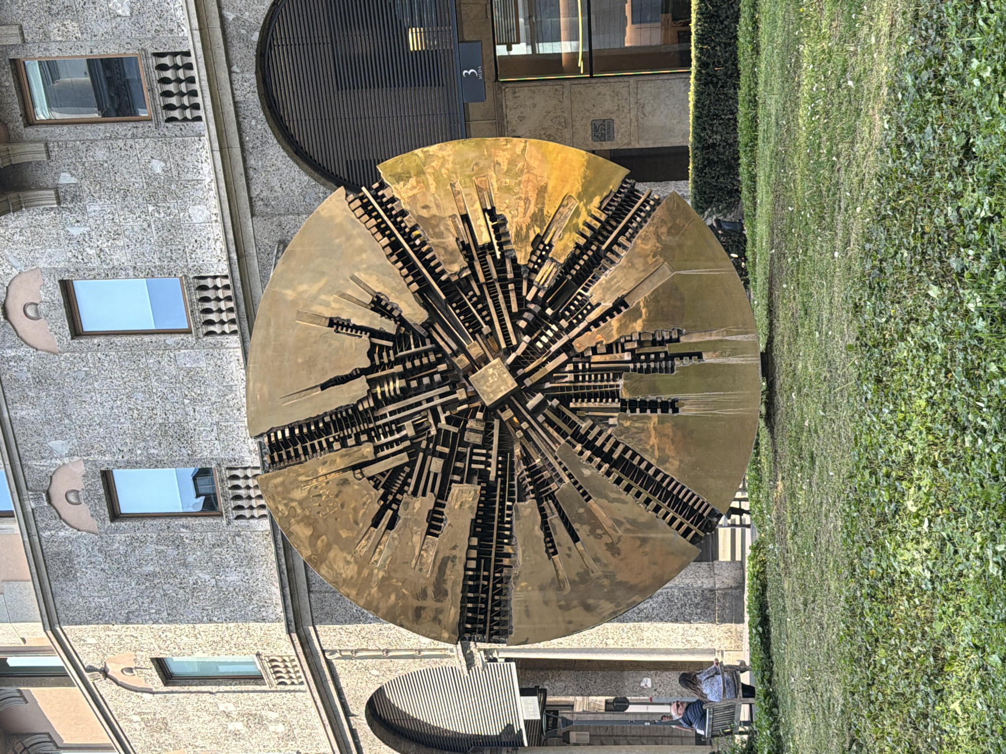 The image shows the sculpture "Sfera Grande" by Arnaldo Pomodoro in Milan, Italy. The sculpture is a large, bronze sphere with a complex, fractured surface, positioned on a stone platform near a building. The building is multi-storied with stone cladding and windows. A narrow green hedge separates the sculpture from a grassy area. Two people are seated on a bench in the foreground, and another person is standing nearby.