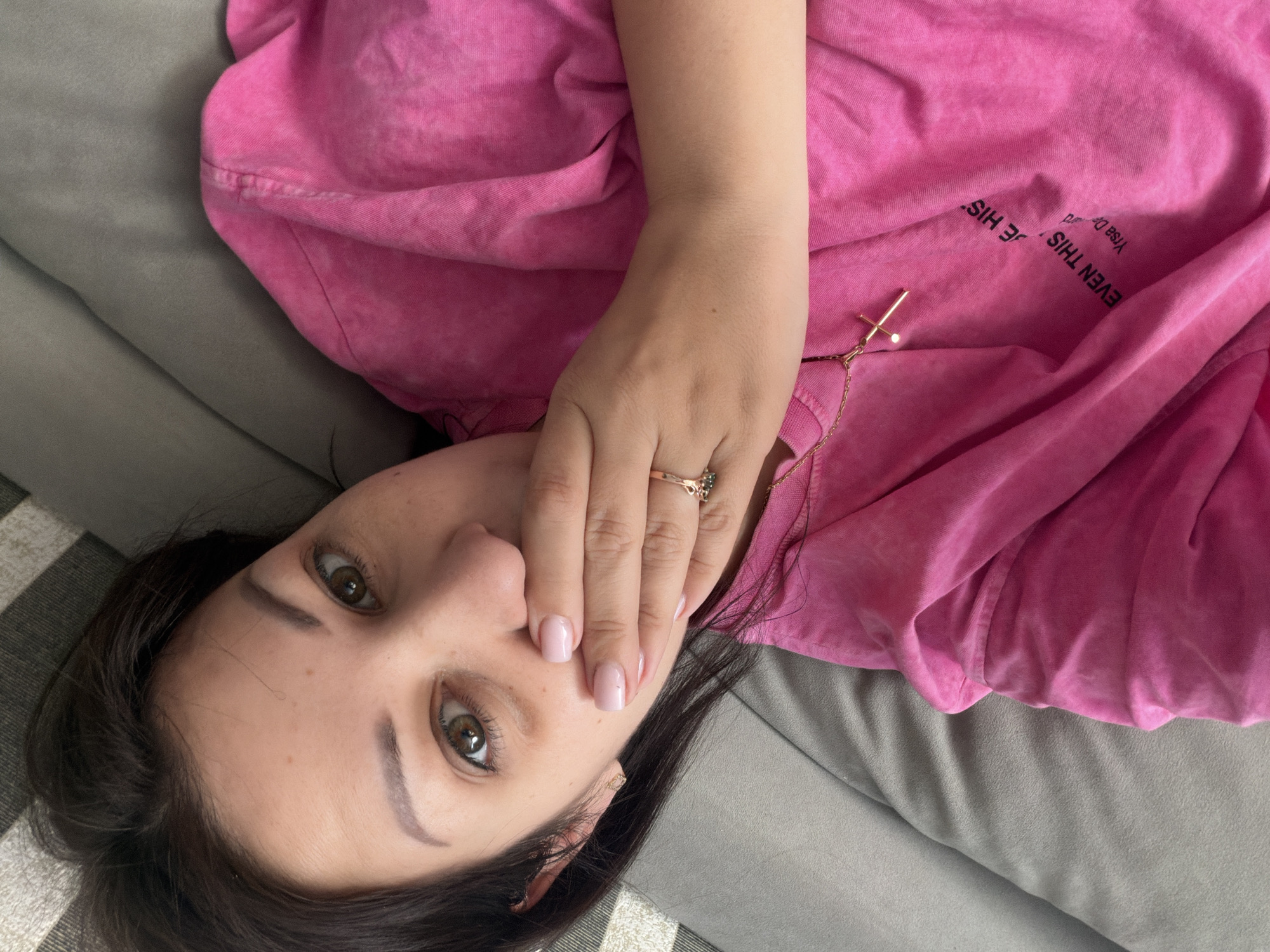 A woman lies on a grey couch, covering her mouth with her right hand. She wears a pink t-shirt with a gold cross necklace. Her left hand is visible on her chest, adorned with a ring. The t-shirt has faded black text that reads "EVEN THIS" and "Yrsa De". The woman has dark brown hair and brown eyes. The background consists of the grey couch and a striped cushion. The image is taken from a high angle. The location is not specified in the image.