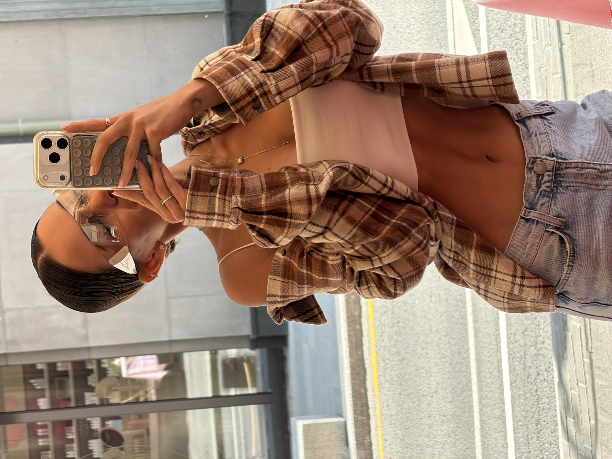 A young woman is captured taking a reflection selfie in what appears to be a large glass window, with an urban outdoor setting reflected behind her. She is dressed casually in an open brown and beige plaid long-sleeve shirt worn over a white crop top, revealing her midriff and belly button. Light-wash denim jeans complete her outfit.
She holds a light-colored smartphone with a clear case that features a grey grid of suction cups on its back. Her hands are adorned with several rings, and a delicate silver necklace with small charms or pendants hangs around her neck. A small crescent moon tattoo is visible on her left wrist. Her dark hair is pulled back, and she wears clear-framed glasses, a stud earring, and another ring-like earring in her right ear. Her fingernails are neatly manicured with a light polish.
The reflected background shows grey concrete surfaces, likely a sidewalk or street, and reflections of buildings, indicating an urban environment. Bright sunlight illuminates the scene, suggesting it is daytime. No visible text is present in the image. The setting is in Moscow, Russia.