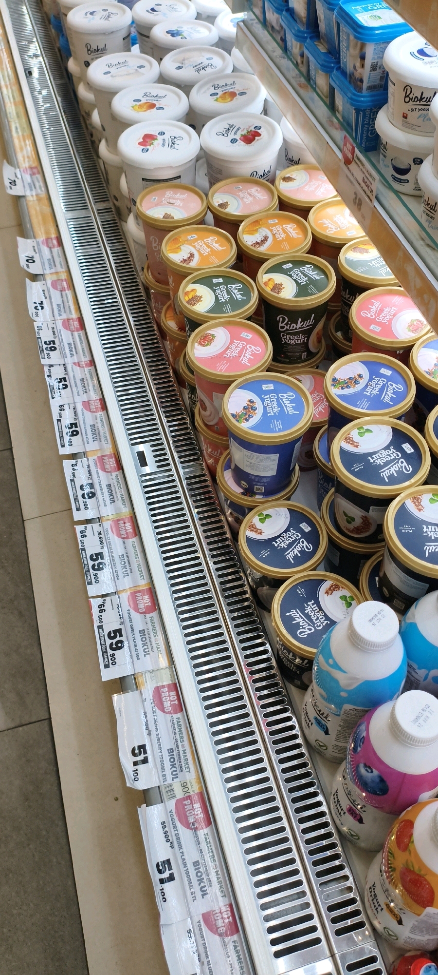This is a close-up, eye-level shot of a refrigerated display case filled with various yogurt products at a Farmers Market in West Jakarta, Indonesia. The dominant items are tubs and bottles of "Biokul" brand yogurt, with labels indicating flavors like "Greek Yogurt," "Plain," "Strawberry," "Blueberry," and "Mango." The display is meticulously organized, with rows of yogurt tubs stacked on shelves. Some tubs have illustrated lids depicting fruit, while others have simple white lids with the brand logo. Several promotional tags are visible, attached to the shelving, advertising "HOT PROMO" prices. The environment appears to be a commercial food retail setting, with a tiled floor visible at the bottom of the frame and the reflective surface of the refrigerated case. No people are visible in the image.